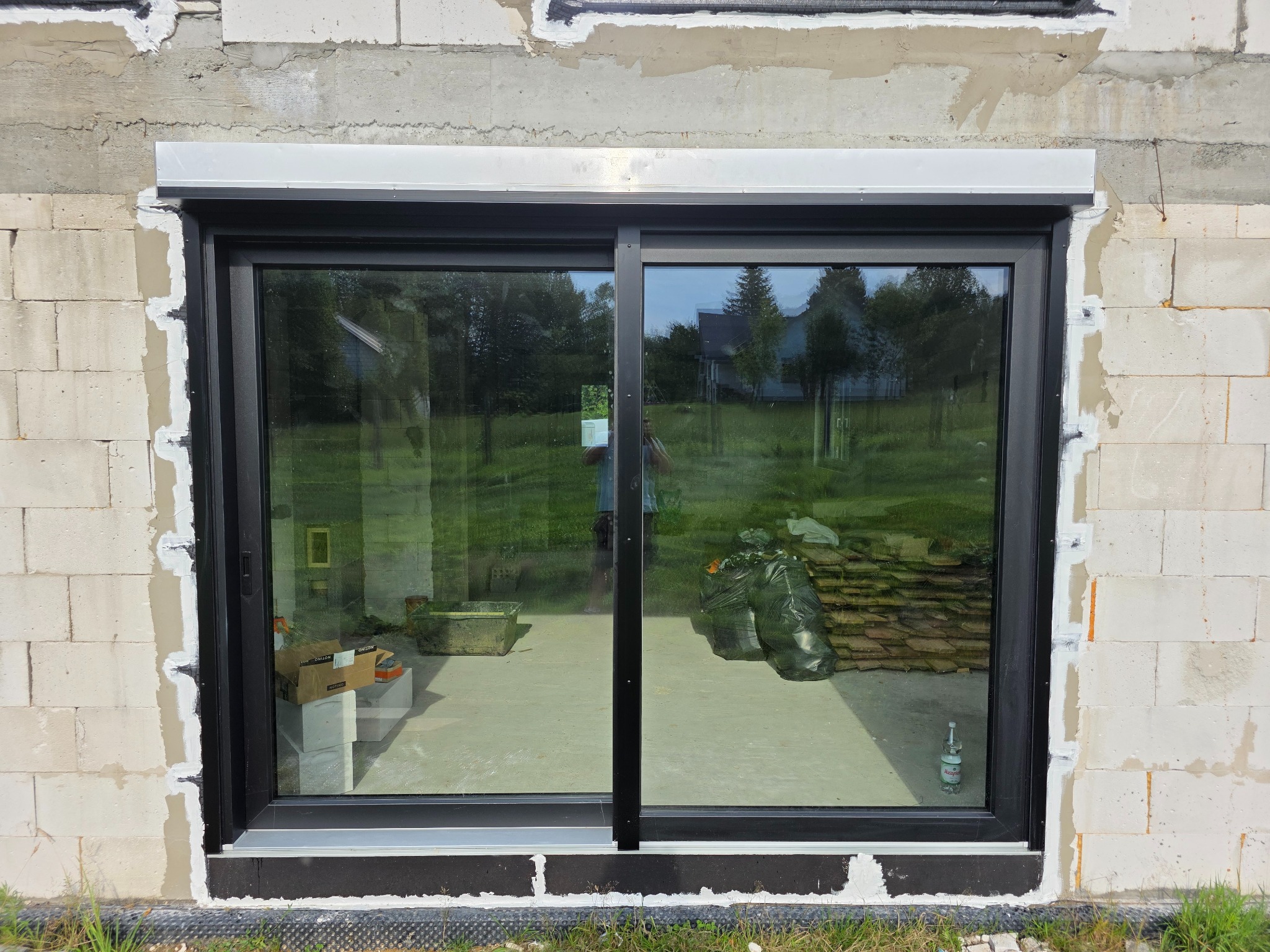 Czarne, przesuwne okno tarasowe z zamontowaną roletą zewnętrzną, widoczne na tle surowej ściany z betonu komórkowego. W tle zieleń trawy i drzew.