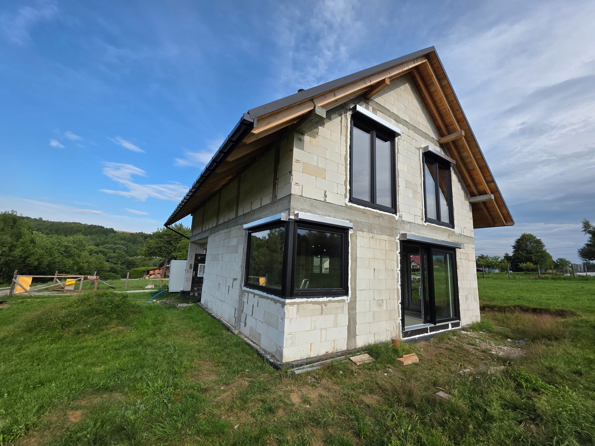 Surowy dom z bloczków z zamontowanymi oknami w czarnych ramach, drewnianą konstrukcją dachu i widokiem na zielone otoczenie. Etap budowy.