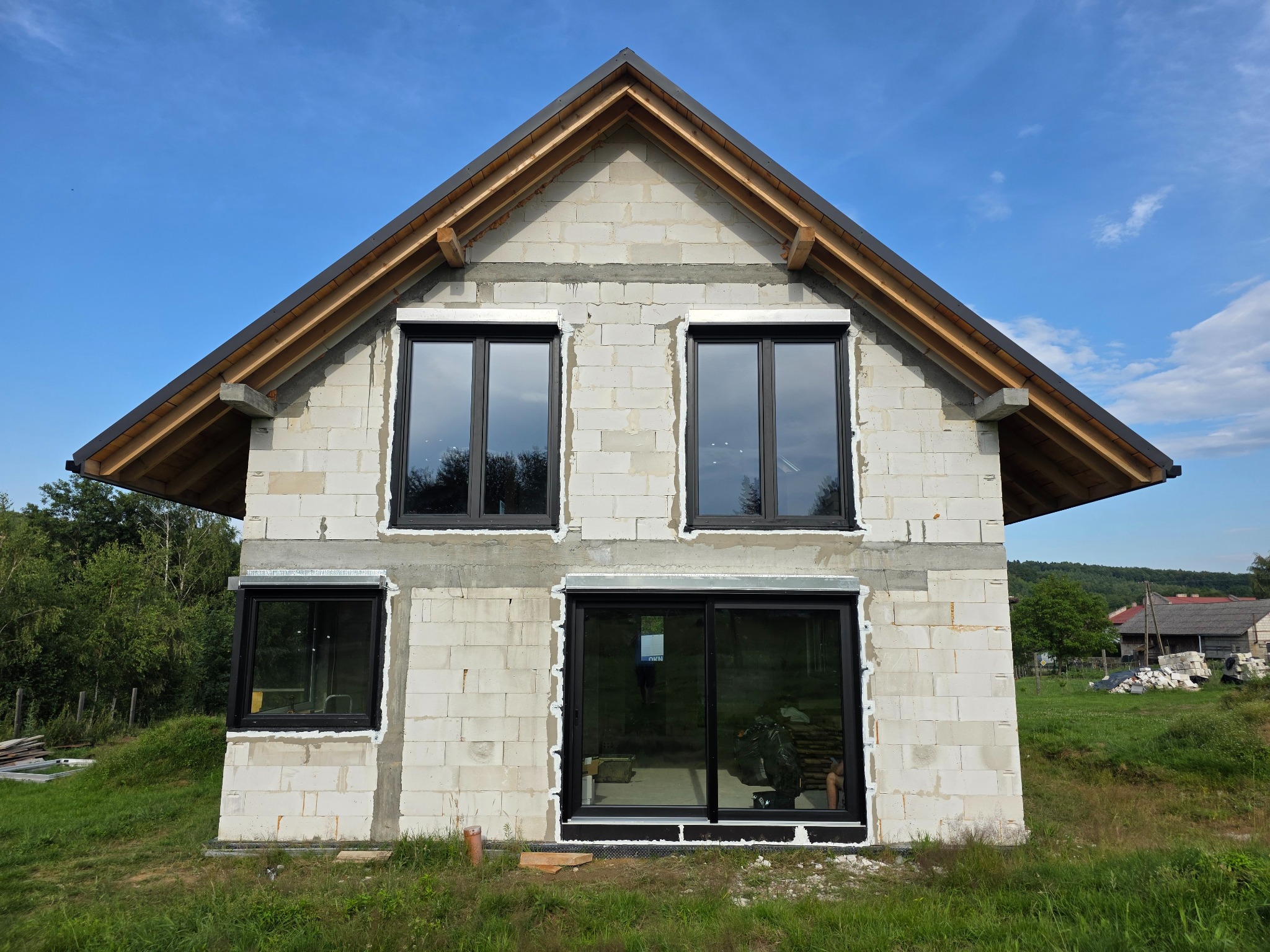 Dom w trakcie budowy z zamontowanymi oknami z czarnymi ramami. Widoczne surowe ściany z białego bloczka i elementy konstrukcji dachu.