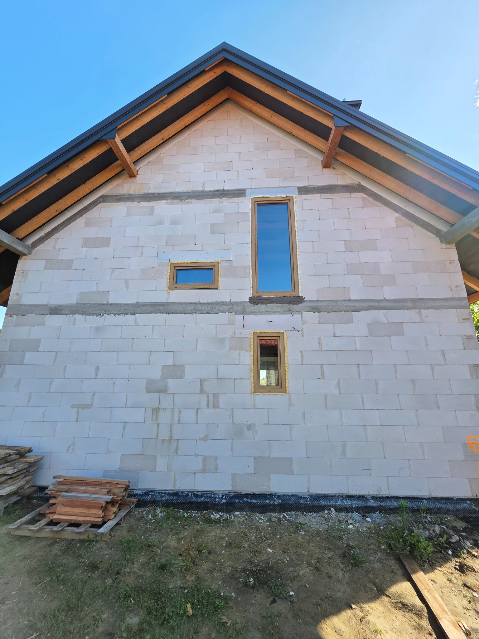 Ściana domu w trakcie budowy z zamontowanymi oknami o drewnianych ramach. Widoczne bale dachowe i niebieskie niebo. Obok leżą drewniane deski.