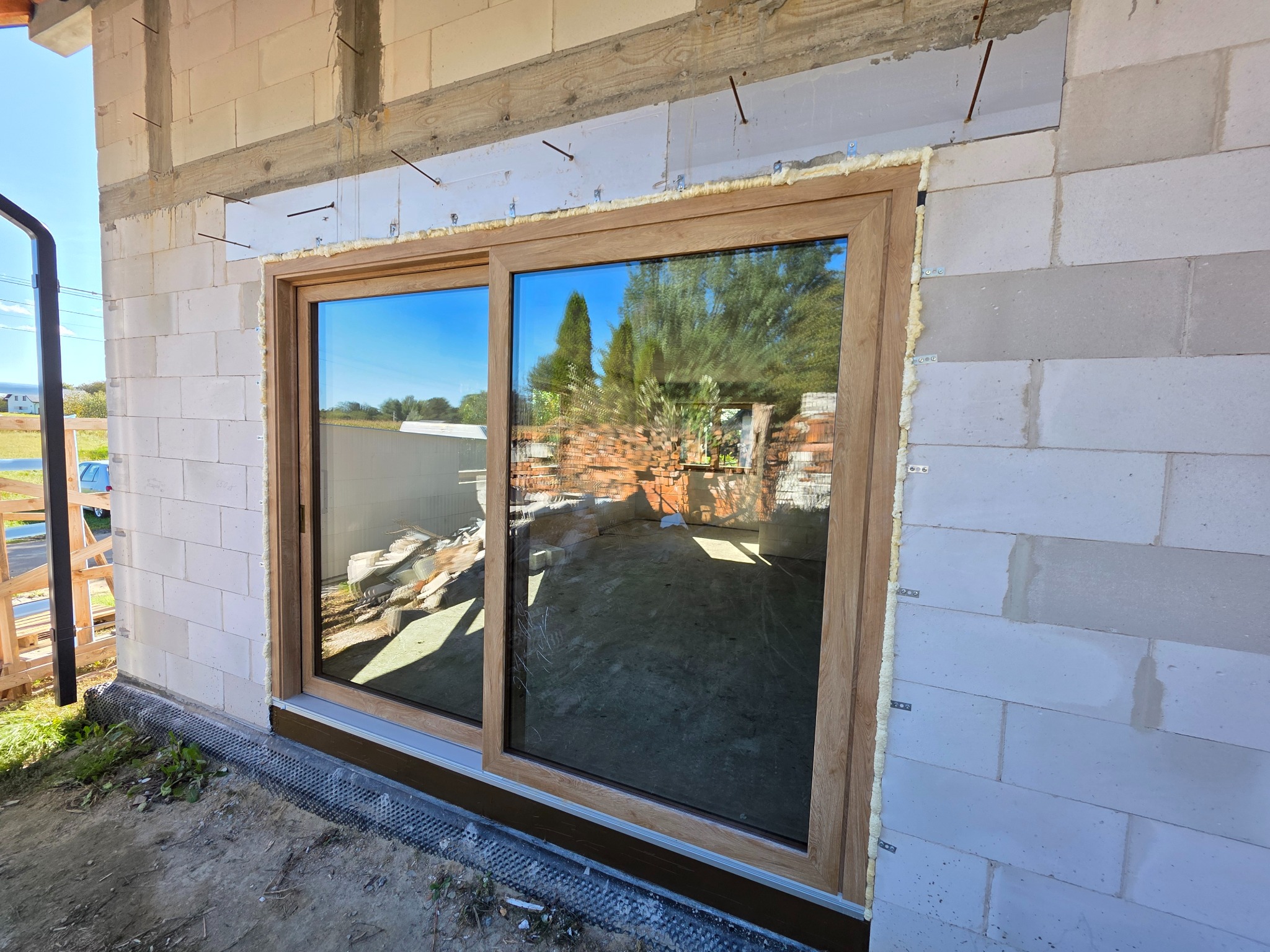 Montaż okna przesuwnego w surowym stanie budynku. Widoczne pianowanie i mocowania. Okno z drewnopodobną ramą odbija krajobraz.