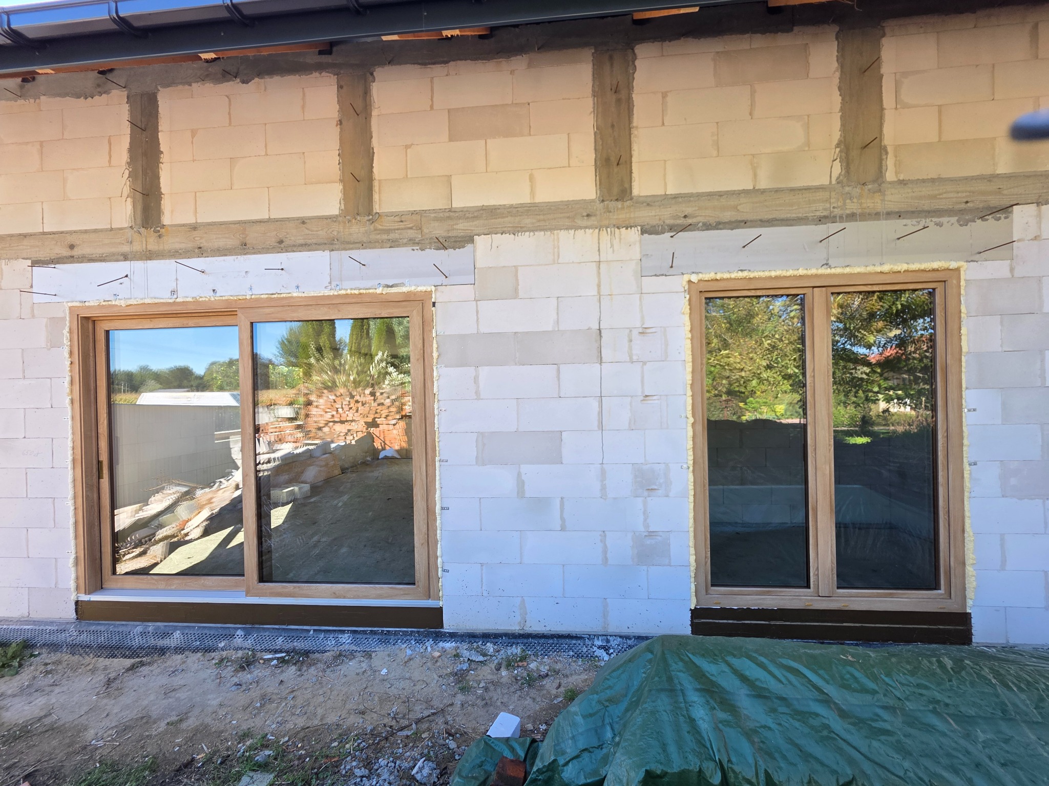 Dwa zamontowane okna z drewnianą ramą w surowej ścianie budynku w trakcie budowy. Widoczne pianka montażowa i elementy konstrukcyjne.