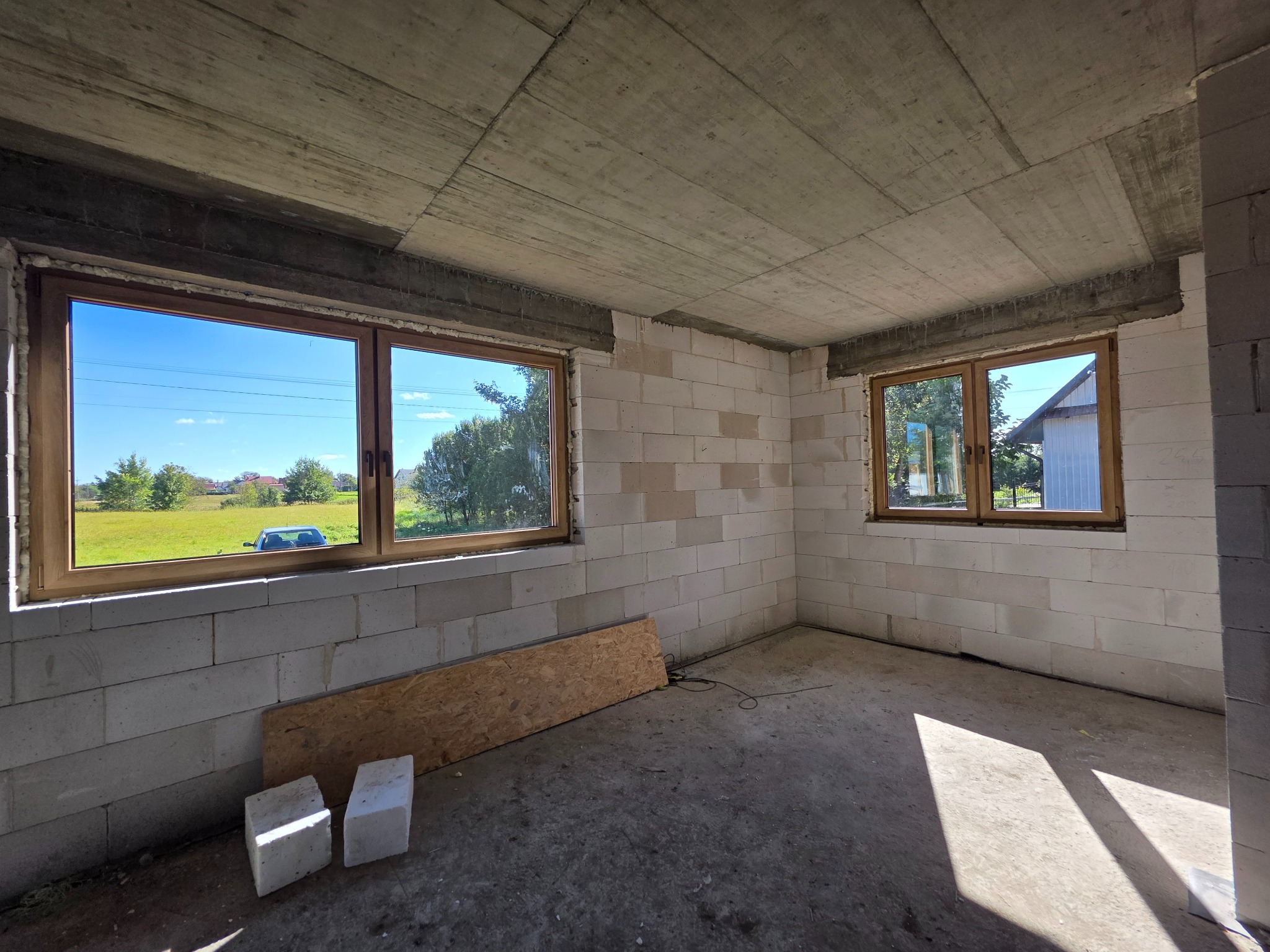 Surowy stan wnętrza z dwoma oknami z drewnianą ramą, widok na zewnątrz zieleń. Ściany z betonu komórkowego, sufit surowy beton. Na podłodze płyta OSB i dwa bloczki.