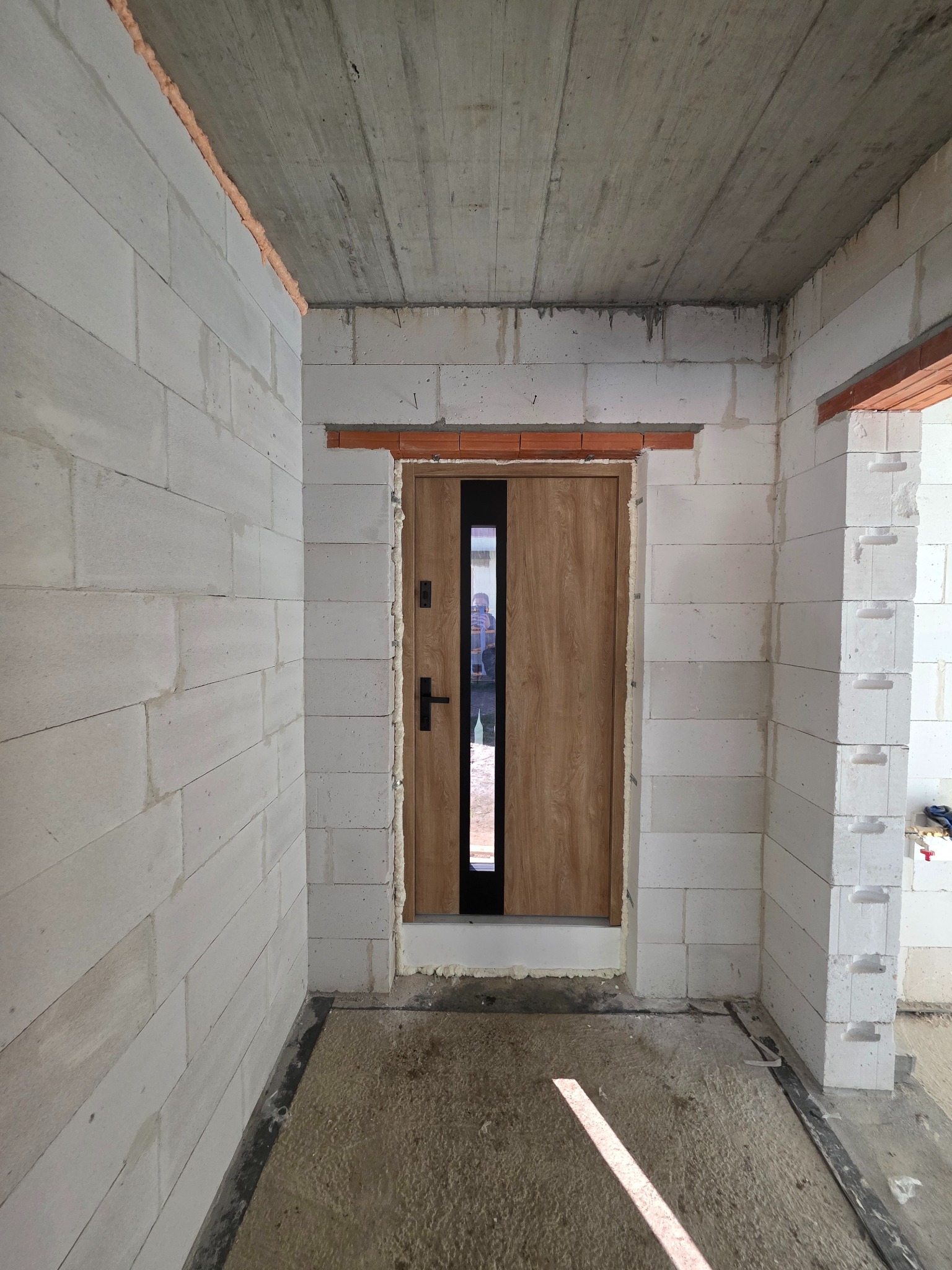 Montaż drzwi wejściowych z szybą w surowym stanie, widoczne białe bloczki i pianka montażowa. Drzwi drewnopodobne z czarnymi akcentami.