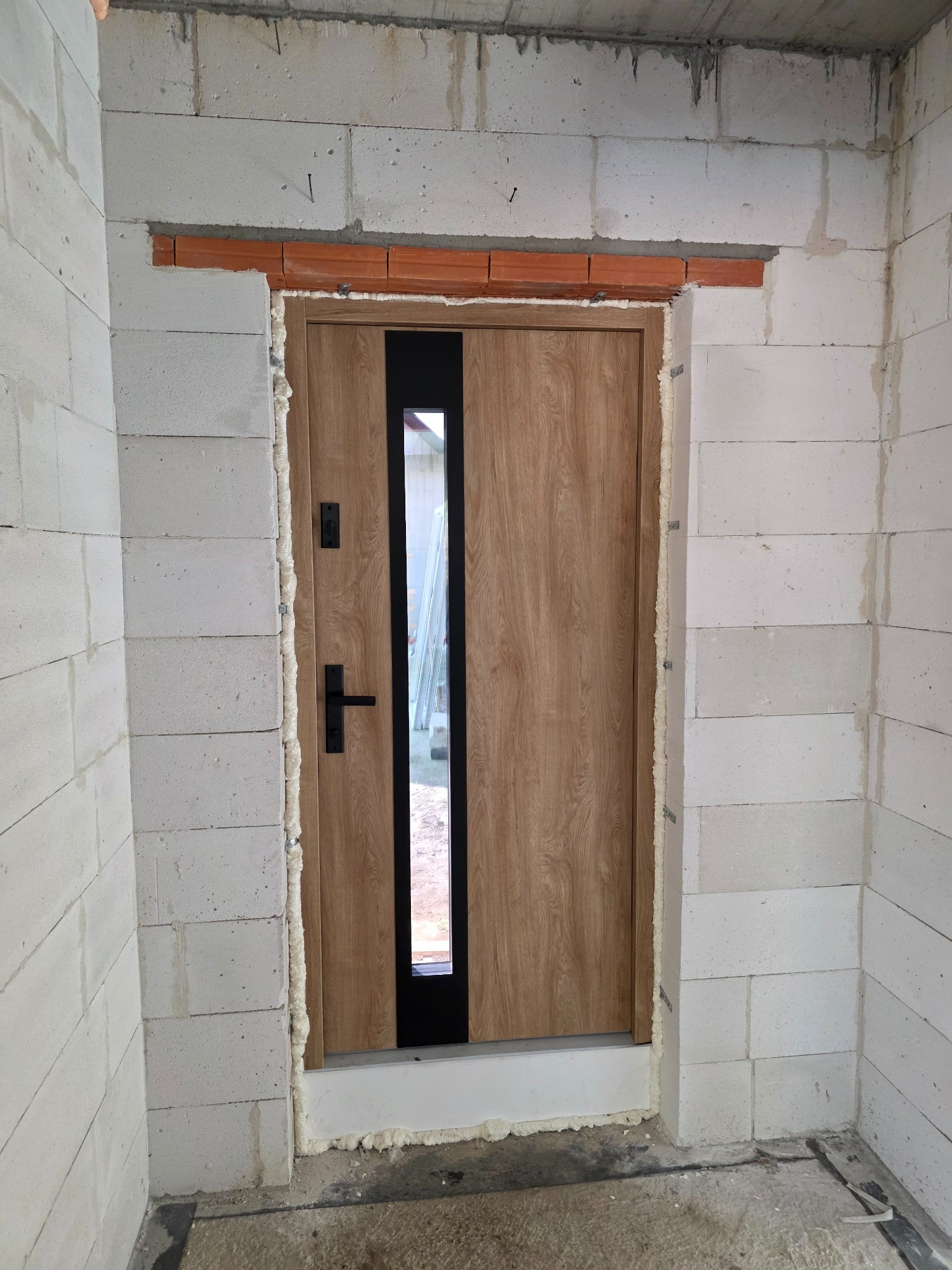 Montaż drzwi wejściowych z szybą w surowym stanie. Widoczne białe bloczki betonowe i pianka montażowa. Czarna klamka i szyba w pionowym pasku.