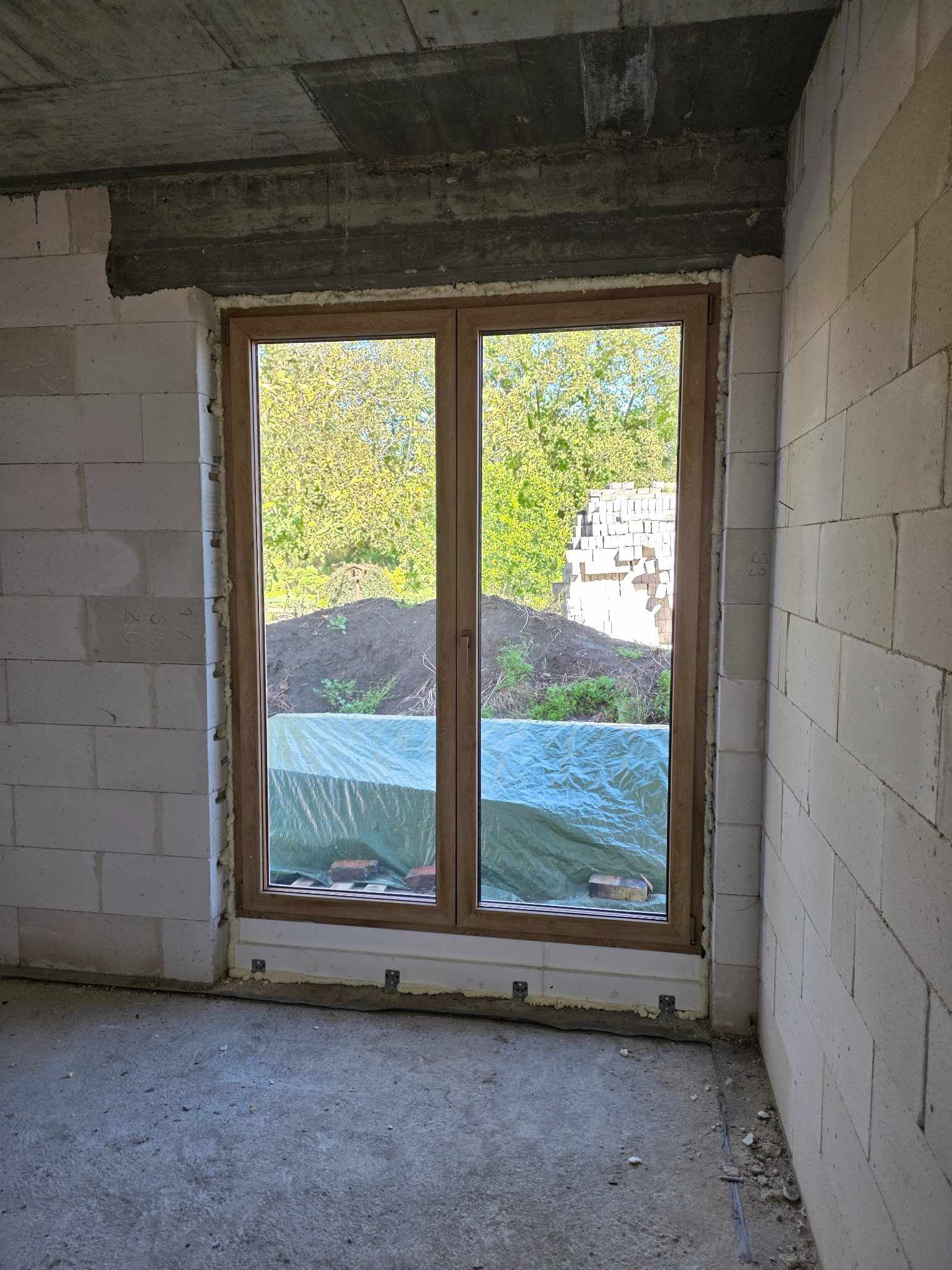 Wnętrze w trakcie budowy: dwuskrzydłowe okno z brązową ramą, osadzone w surowej ścianie z betonu komórkowego, zabezpieczone folią od zewnątrz. Widok na zieleń.