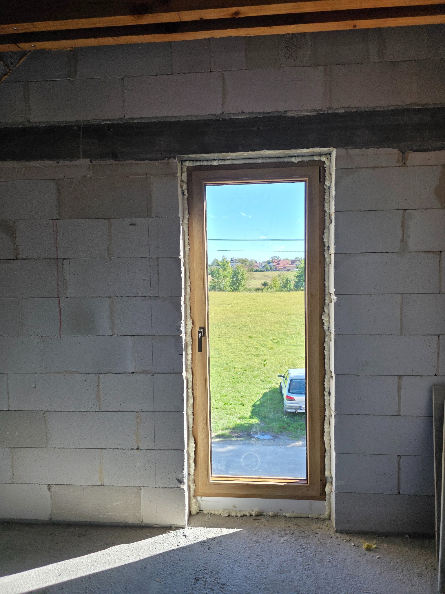 Montaż okna z brązową ramą w surowej ścianie z betonu komórkowego. Widoczne pianowanie uszczelniające. Za oknem zielona łąka i fragment zaparkowanego auta.