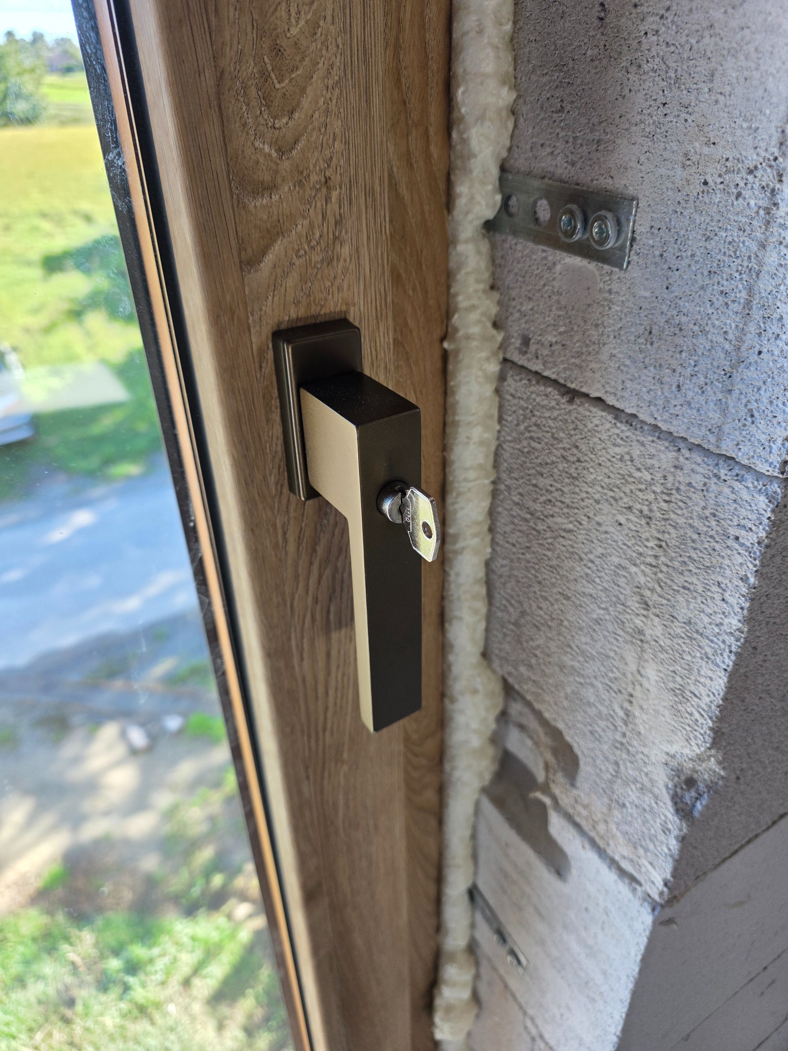 Zamknięte okno z drewnopodobną ramą i czarną klamką, zabezpieczone kluczem. Widoczne mocowanie do muru i pianka montażowa.