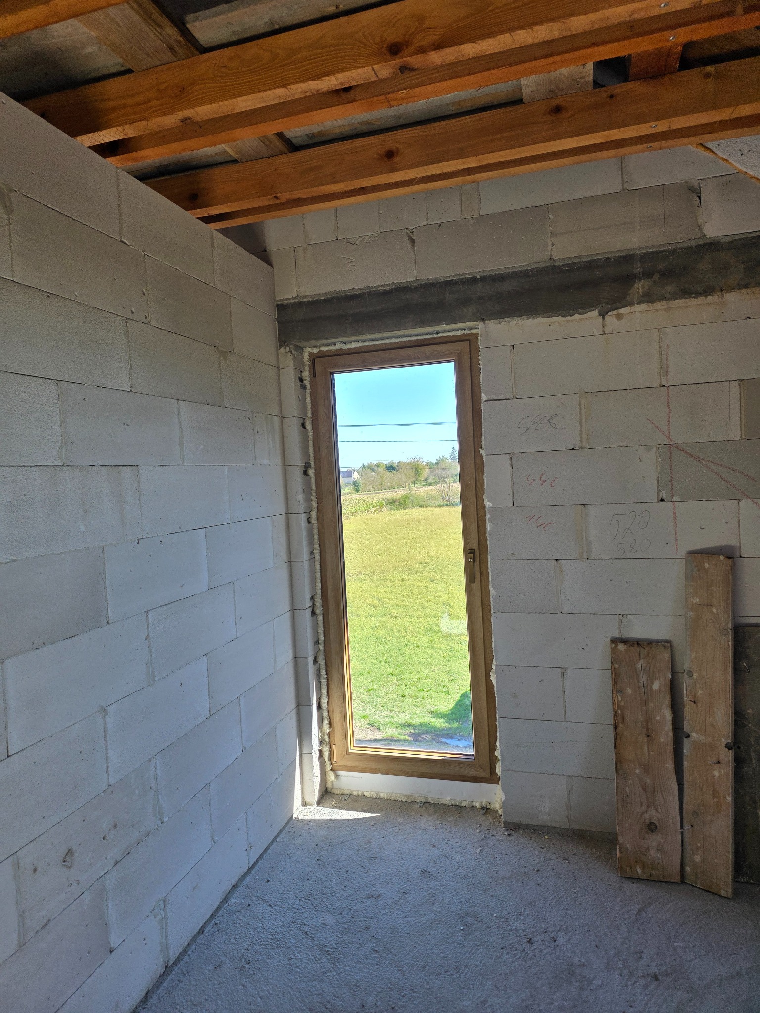 Wnętrze surowego budynku z oknem w drewnianej ramie, widok na zewnątrz na zielone pole. Ściany z białych bloczków, surowy beton na podłodze, drewniane belki stropowe.
