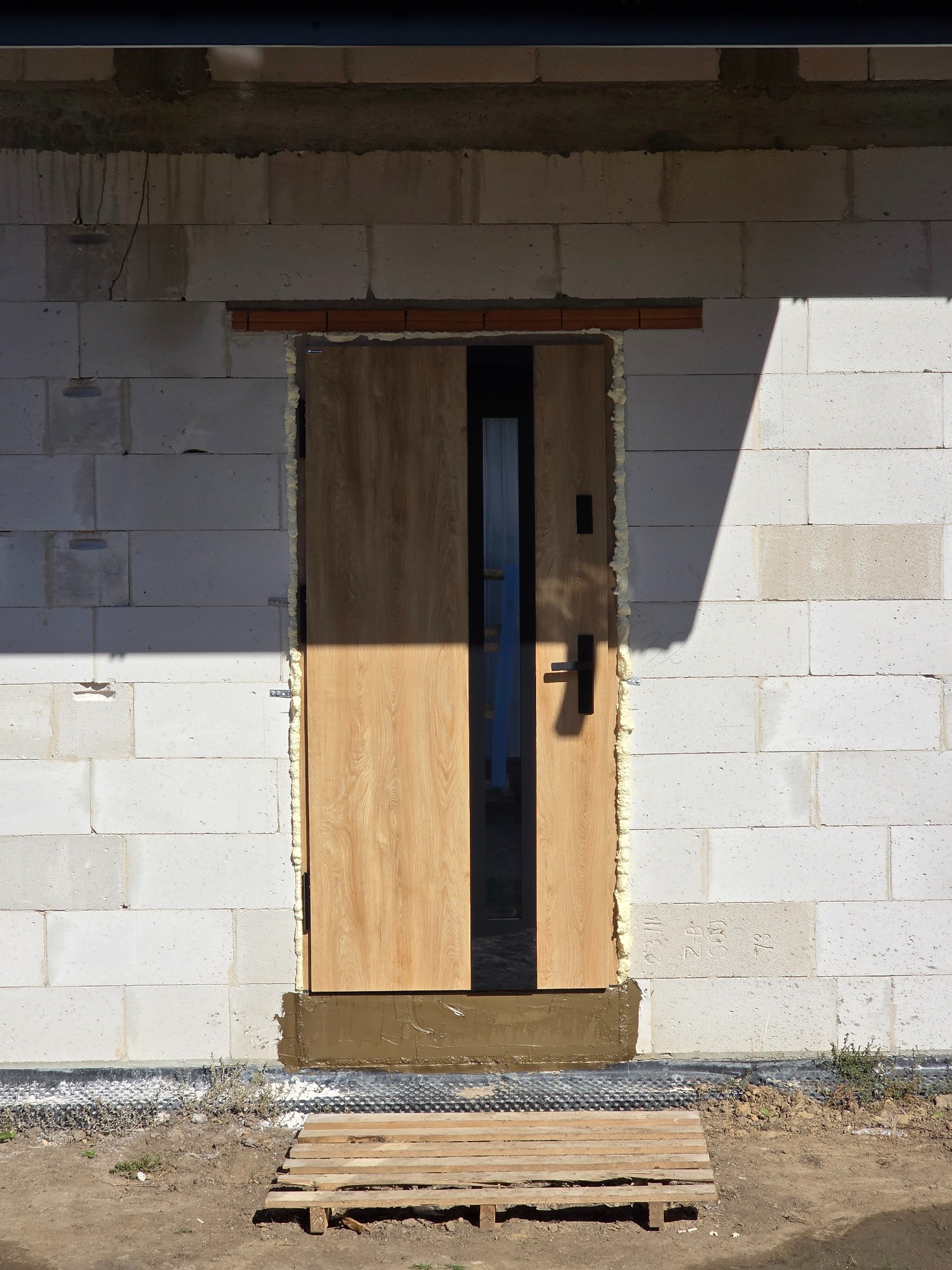 Montaż drzwi zewnętrznych z drewnopodobnym wykończeniem, uszczelnionych pianką montażową, w ścianie z białych bloczków. Widoczna paleta drewniana.
