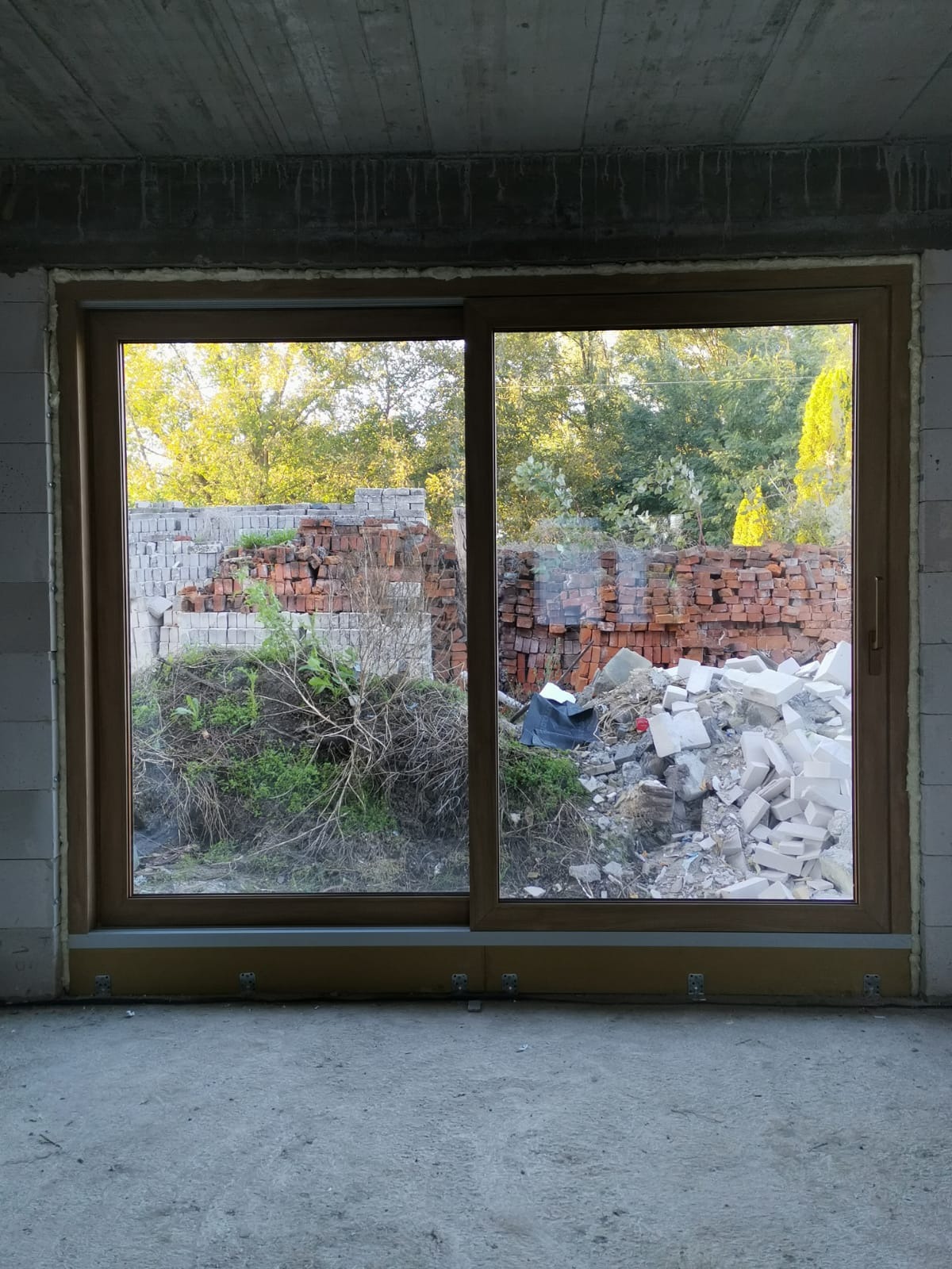 Zamontowane przesuwne okno w surowym stanie, widok na gruz i cegły na zewnątrz. Jasna rama okna, betonowy sufit i ściany z białych bloczków.