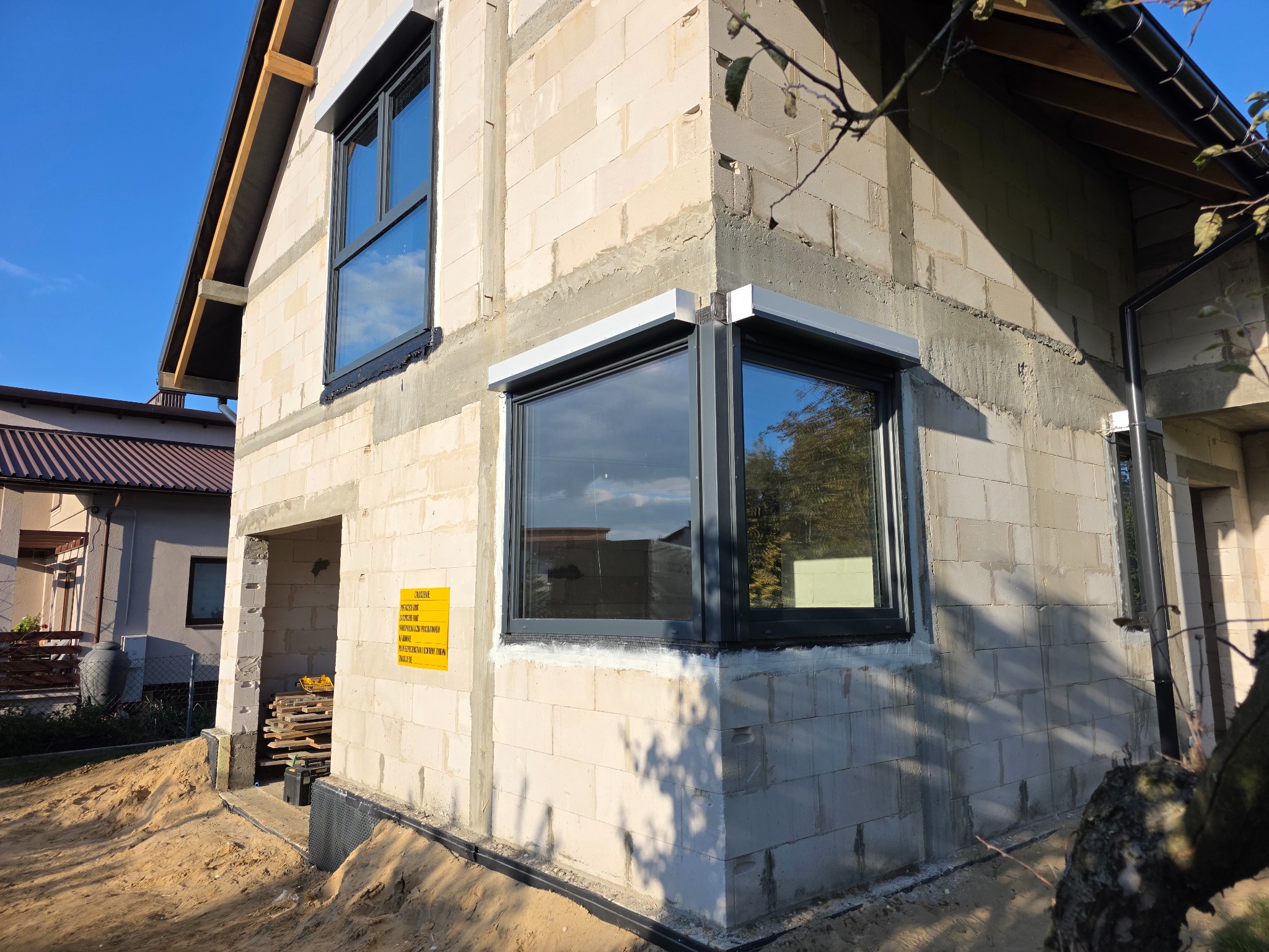 Surowy budynek z szarego betonu komórkowego z zamontowanymi oknami w czarnych ramach. Po prawej stronie widoczna zamontowana roleta zewnętrzna.
