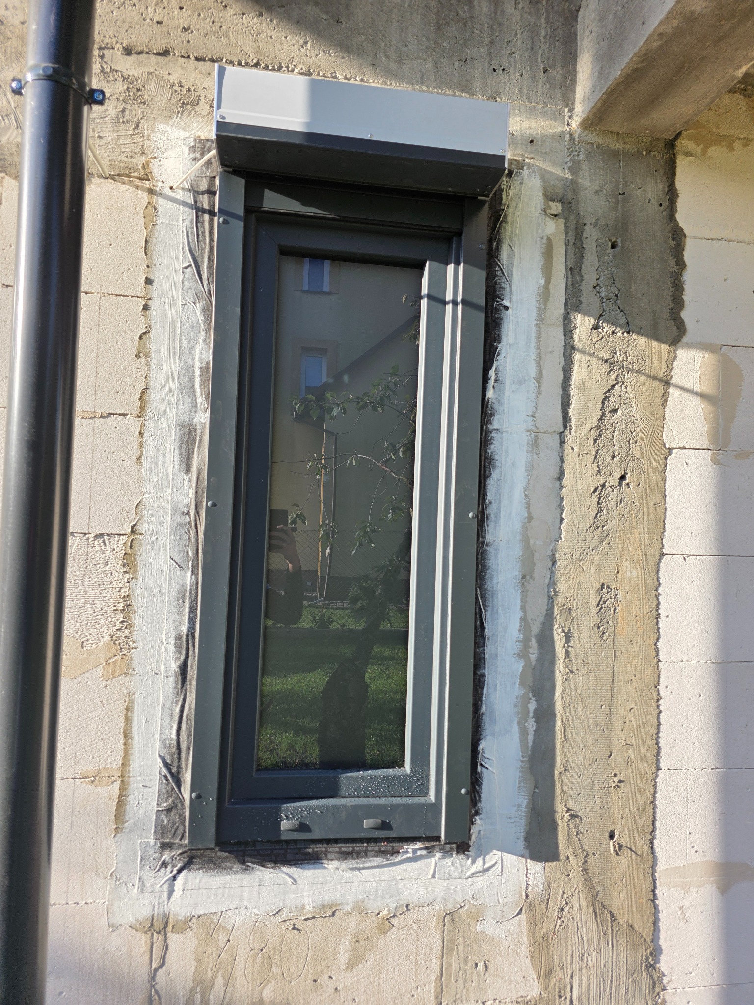 Szare okno z roletą zewnętrzną, zamontowane w surowej ścianie z betonu komórkowego, widoczne ślady uszczelniania. W szybie odbija się budynek i zieleń.