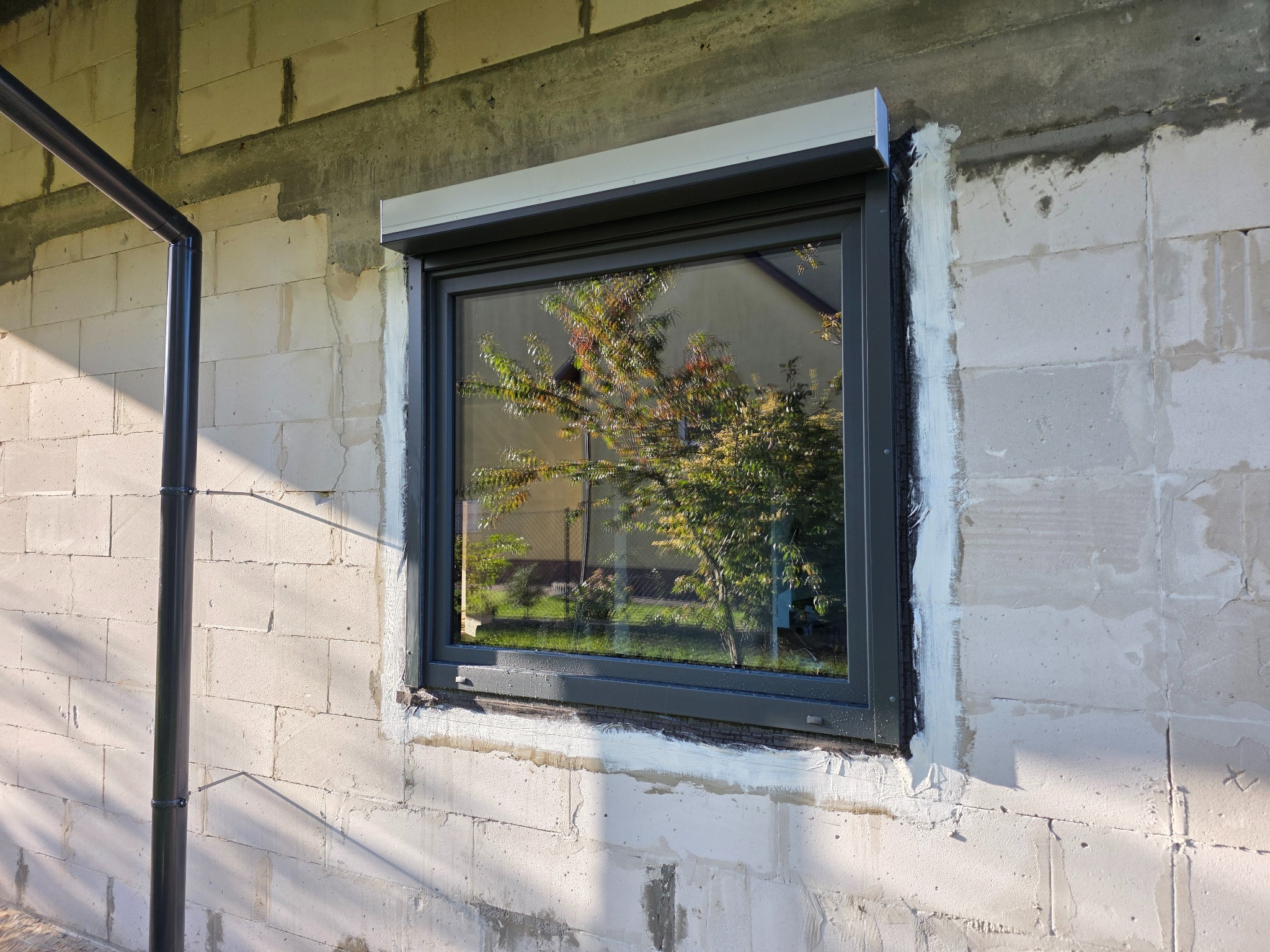 Szare okno z roletą zewnętrzną w surowej ścianie z betonu komórkowego, widoczne ślady po montażu, odbicie drzewa w szybie, rura spustowa obok.