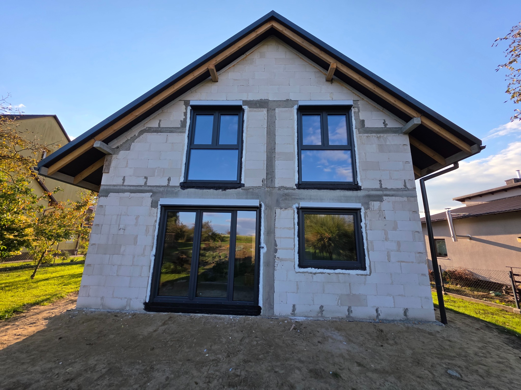 Surowy budynek z zamontowanymi oknami w grafitowej ramie, widoczne refleksy nieba w szybach. Dom w trakcie budowy, ściany z białego bloczka.