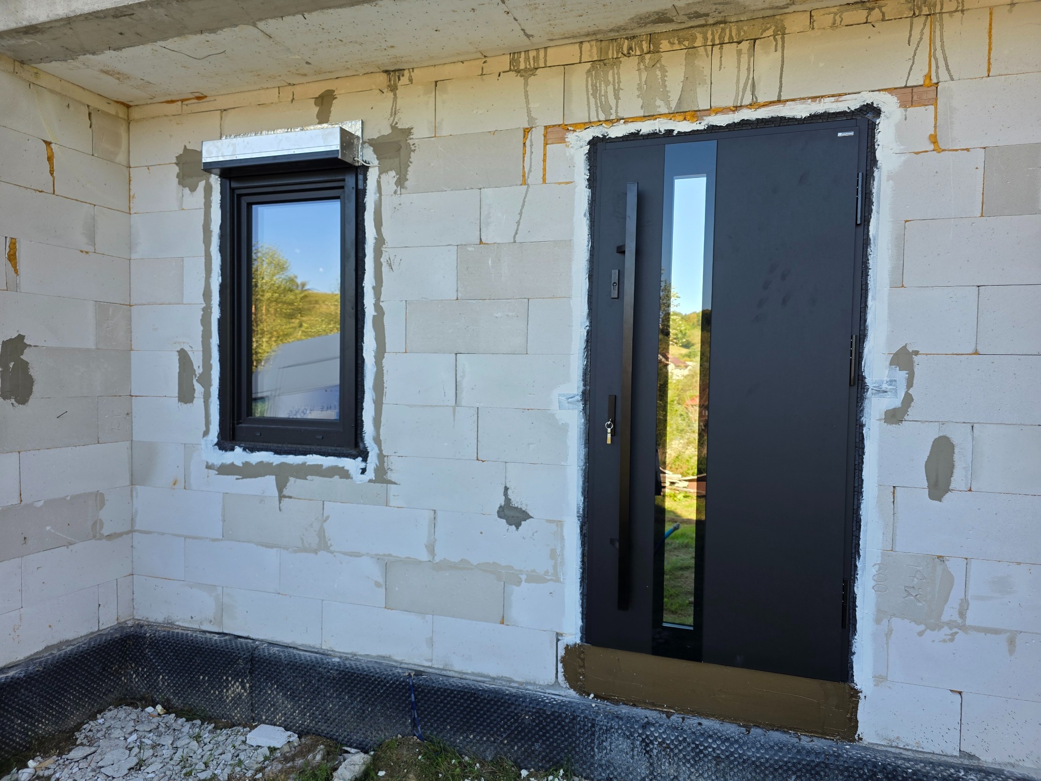 Ciemne drzwi antywłamaniowe z szybą i okno z roletą zewnętrzną w surowym stanie budynku z białego bloczka, widok z zewnątrz.