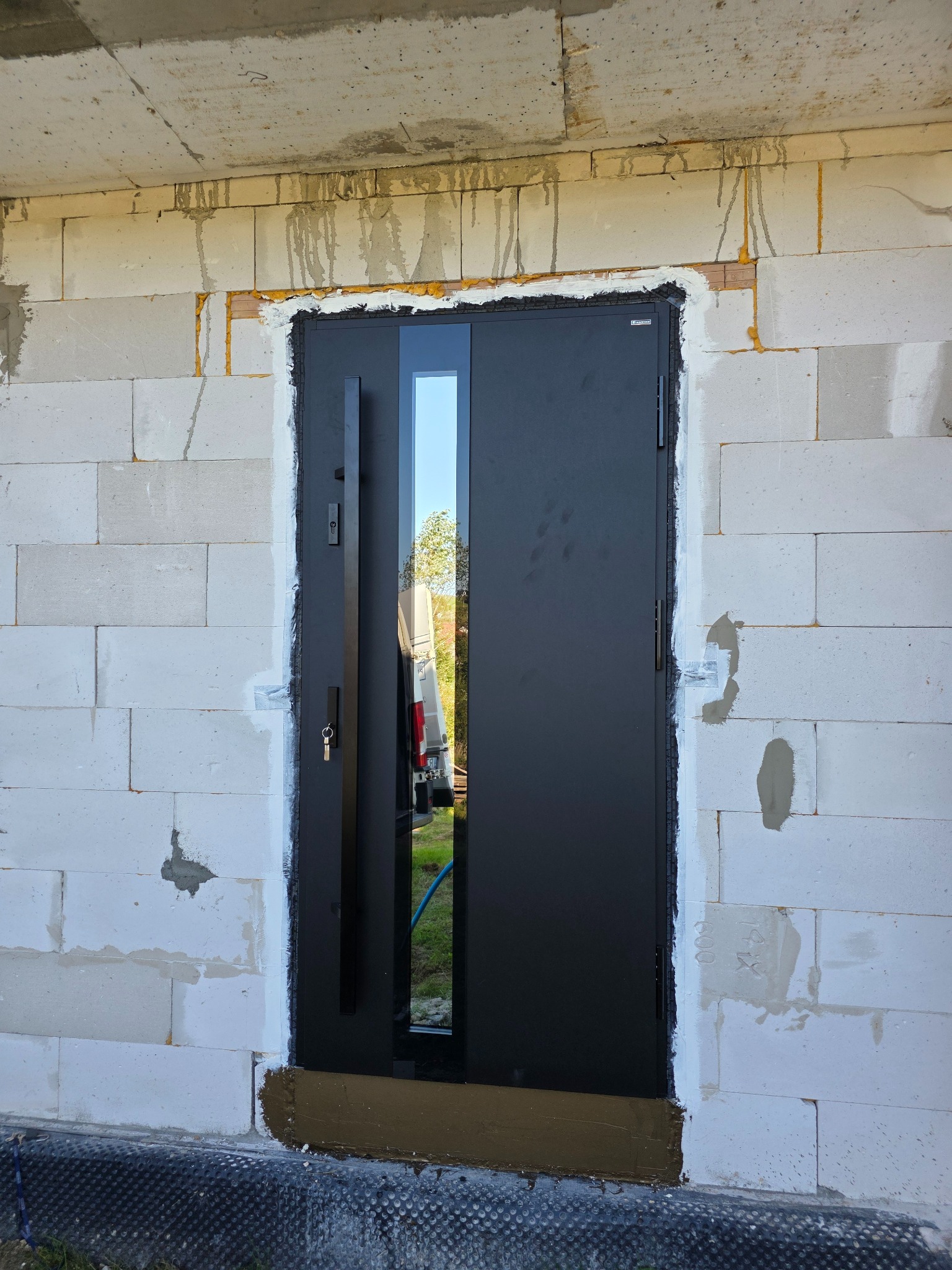 Ciemne drzwi wejściowe z przeszkleniem, zamontowane w surowym murze z betonu komórkowego. Widoczne ślady pianki montażowej i zaprawy. Klucz w zamku.