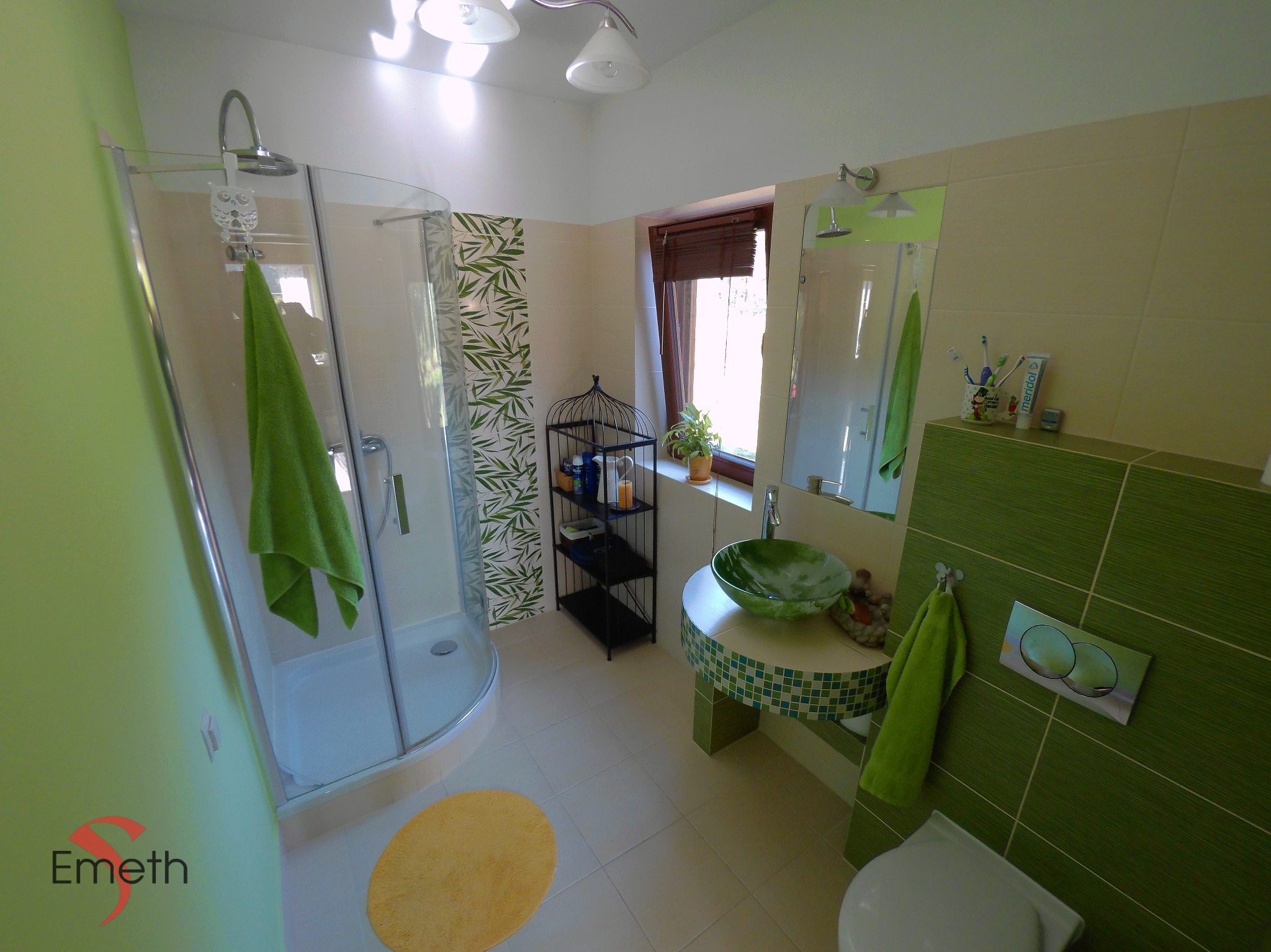 Jasna łazienka z kabiną prysznicową, zieloną umywalką na mozaikowym blacie, zielonymi akcentami na ścianach i dodatkach, okrągły żółty dywanik na podłodze z płytek.