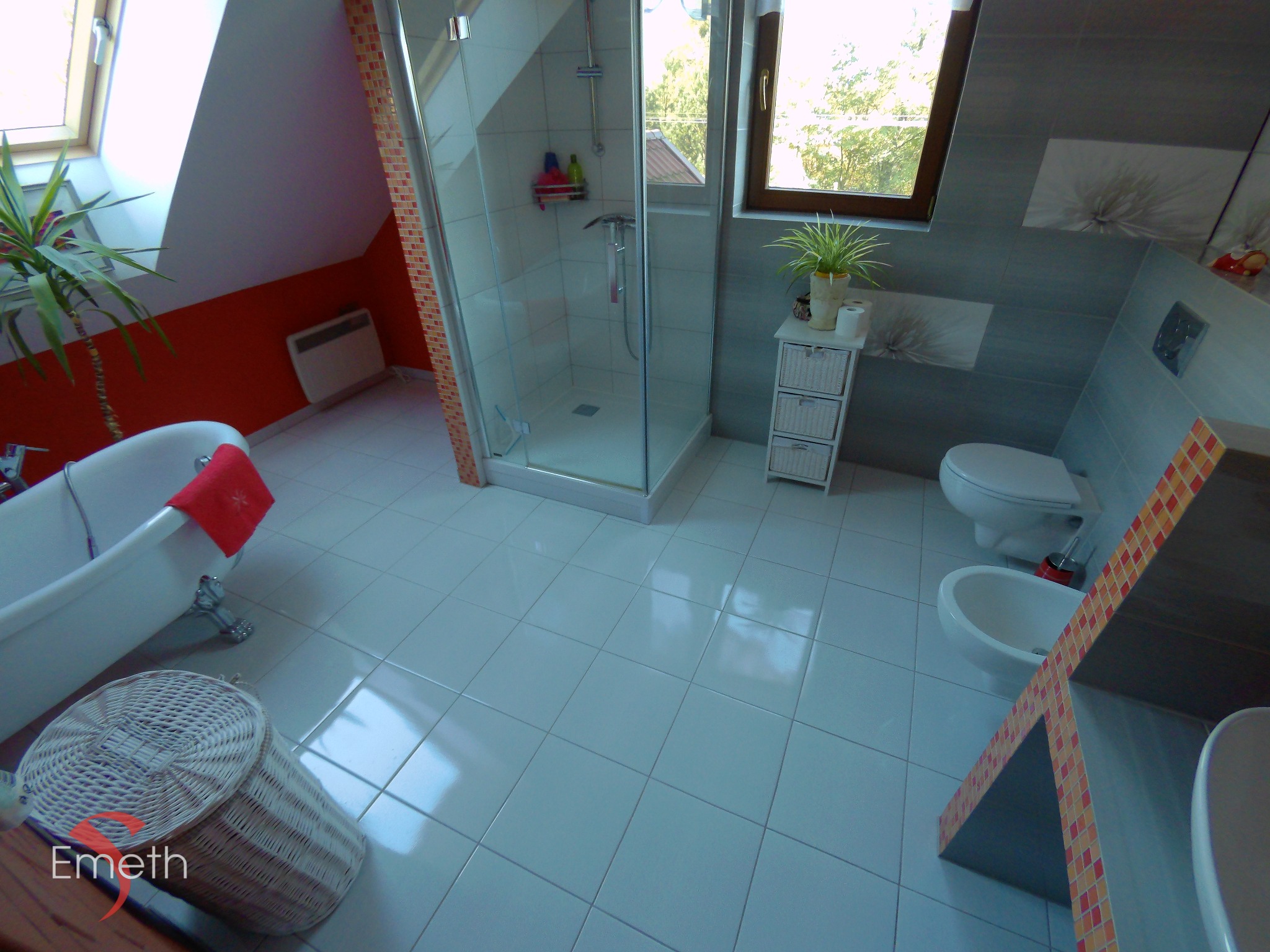 Jasna łazienka na poddaszu z wolnostojącą wanną, kabiną prysznicową z mozaiką, bidetem i toaletą, doświetlona oknem dachowym i ozdobiona roślinami doniczkowymi oraz grafiką na szarej ścianie.