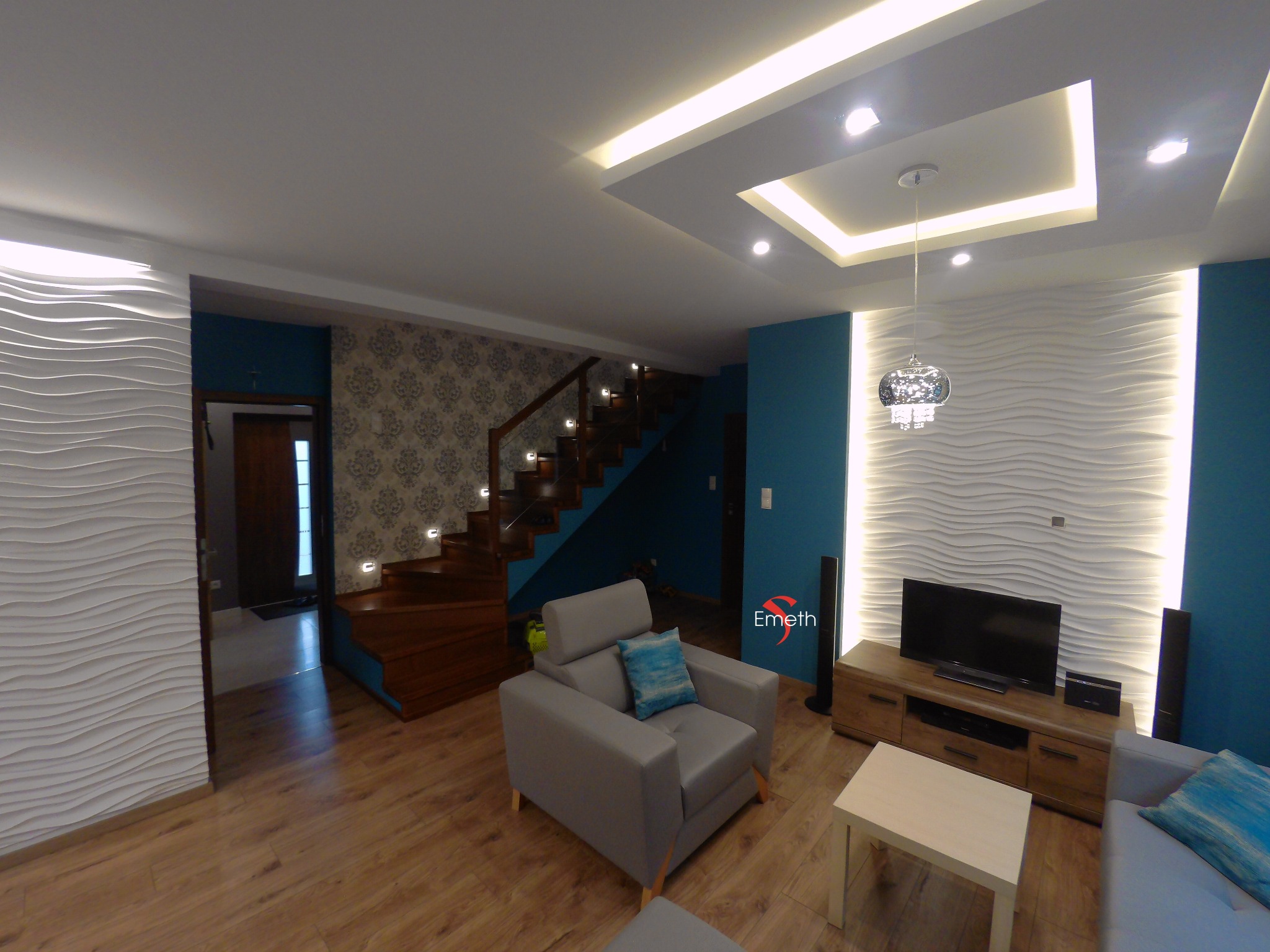 Salon z drewnianymi schodami, szarą sofą z turkusową poduszką, białą ścianą z falistym wzorem i podwieszanym sufitem z oświetleniem LED.