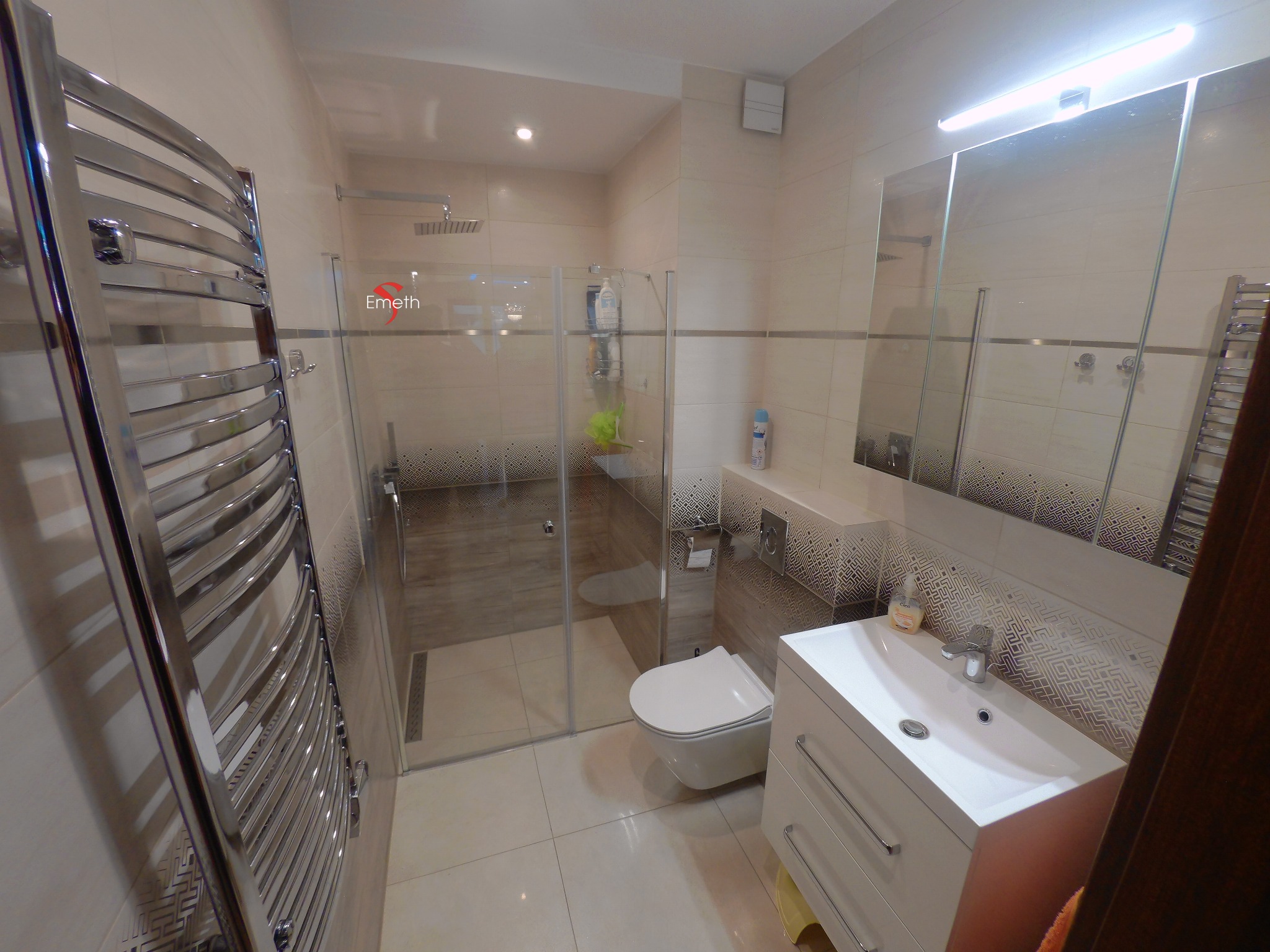 Jasna łazienka po remoncie z kabiną prysznicową typu walk-in, wiszącą toaletą, umywalką z szafką i grzejnikiem drabinkowym, wykończona płytkami z geometrycznym wzorem.