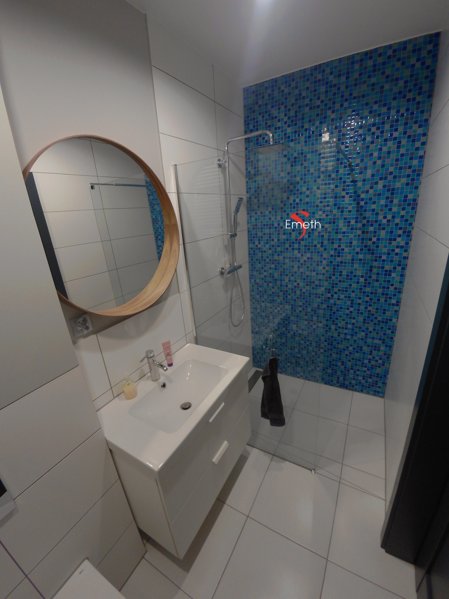 Nowoczesna łazienka z umywalką na szafce, okrągłym lustrem w drewnianej ramie i kabiną prysznicową wyłożoną mozaiką w odcieniach błękitu.