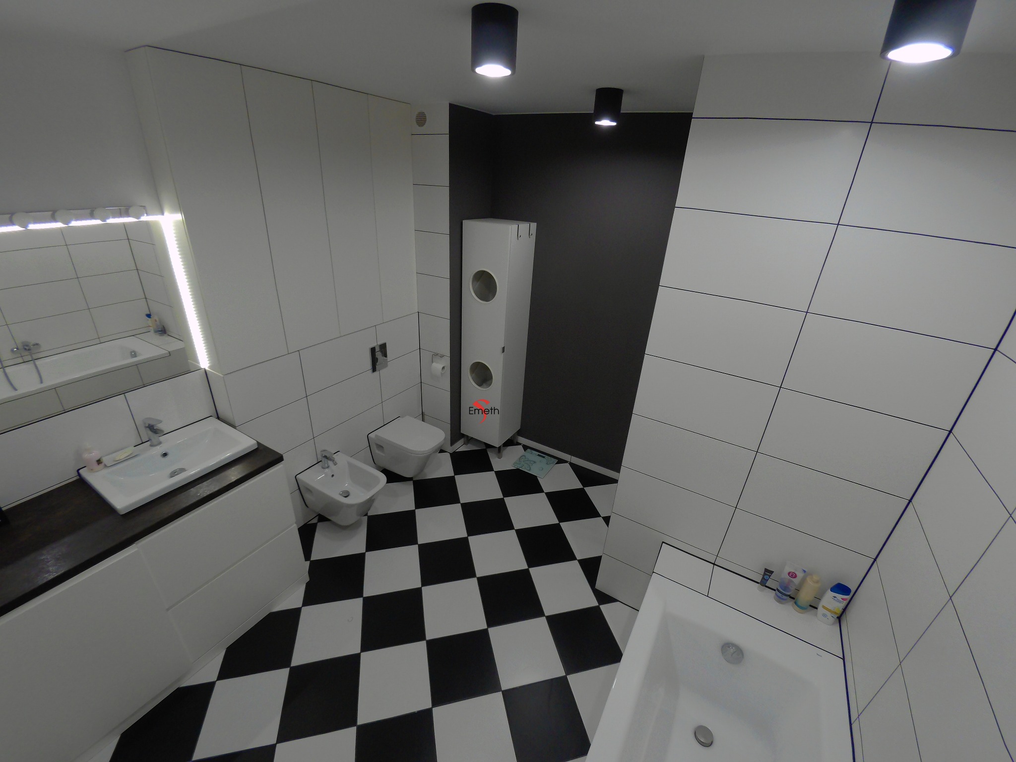 Nowoczesna łazienka z białą armaturą, czarno-białą szachownicą na podłodze, białymi płytkami z granatową fugą i czarną ścianą w tle, oświetlona punktowymi lampami sufitowymi.