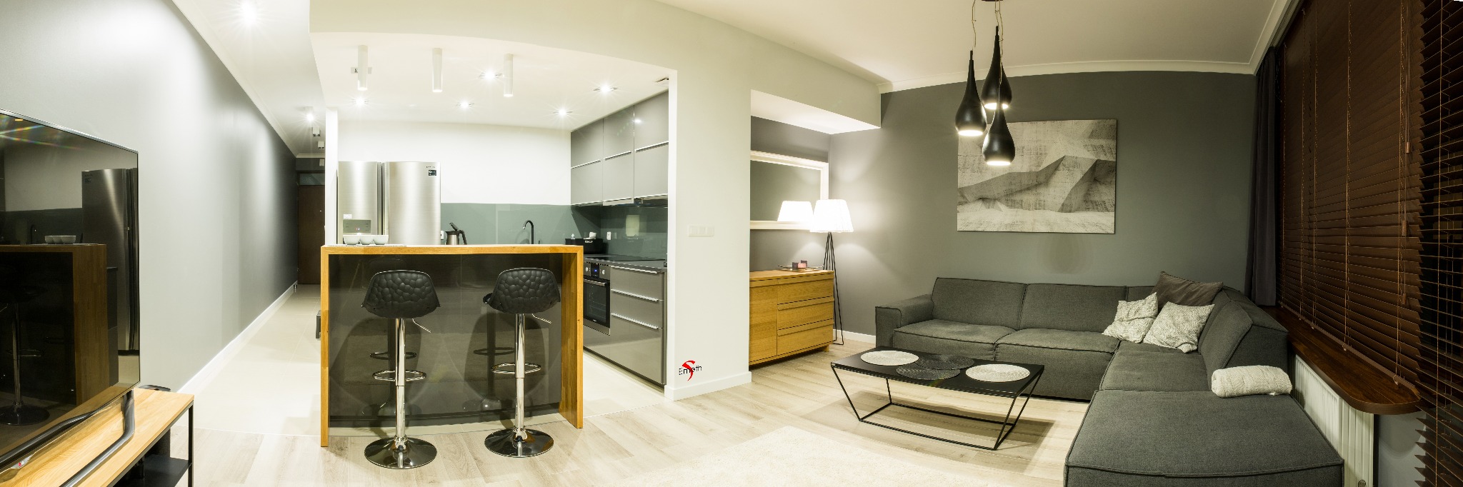 Panoramiczny widok na nowoczesne wnętrze mieszkania z otwartą kuchnią, barem z dwoma stołkami, szarą sofą narożną i drewnianymi żaluzjami.
