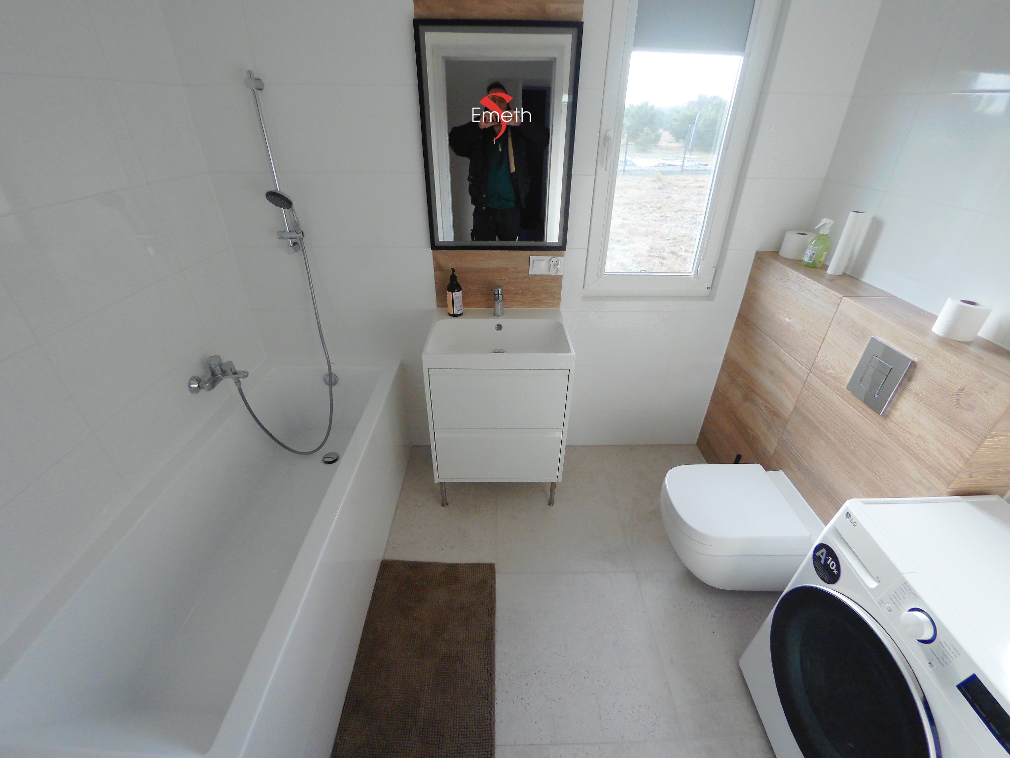 Nowoczesna łazienka z białą armaturą, wanną, umywalką z szafką, toaletą podwieszaną i pralką LG, wykończona białymi płytkami i panelami drewnopodobnymi, z odbiciem osoby w lustrze.