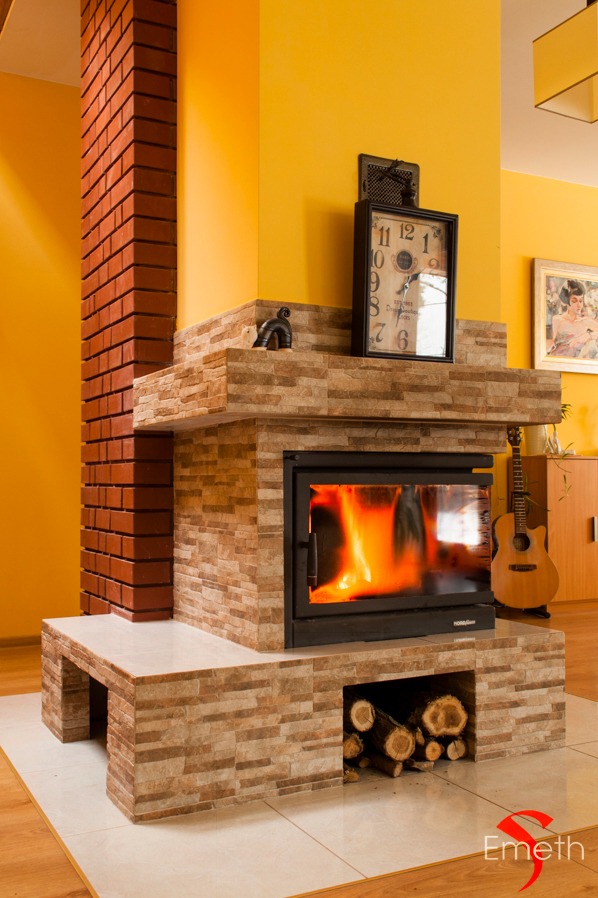 Nowoczesny kominek obłożony kamieniem dekoracyjnym z paleniskiem i zapasem drewna, ceglana kolumna po lewej, zegar i gitara w tle, żółta ściana.