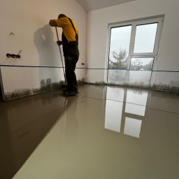 Robert Hodyjas MHR - Pracownik w żółtej koszulce rozprowadza wylewkę samopoziomującą w jasnym pomieszczeniu, zabezpieczone ściany folią, widoczne okno z widokiem na drzewa.