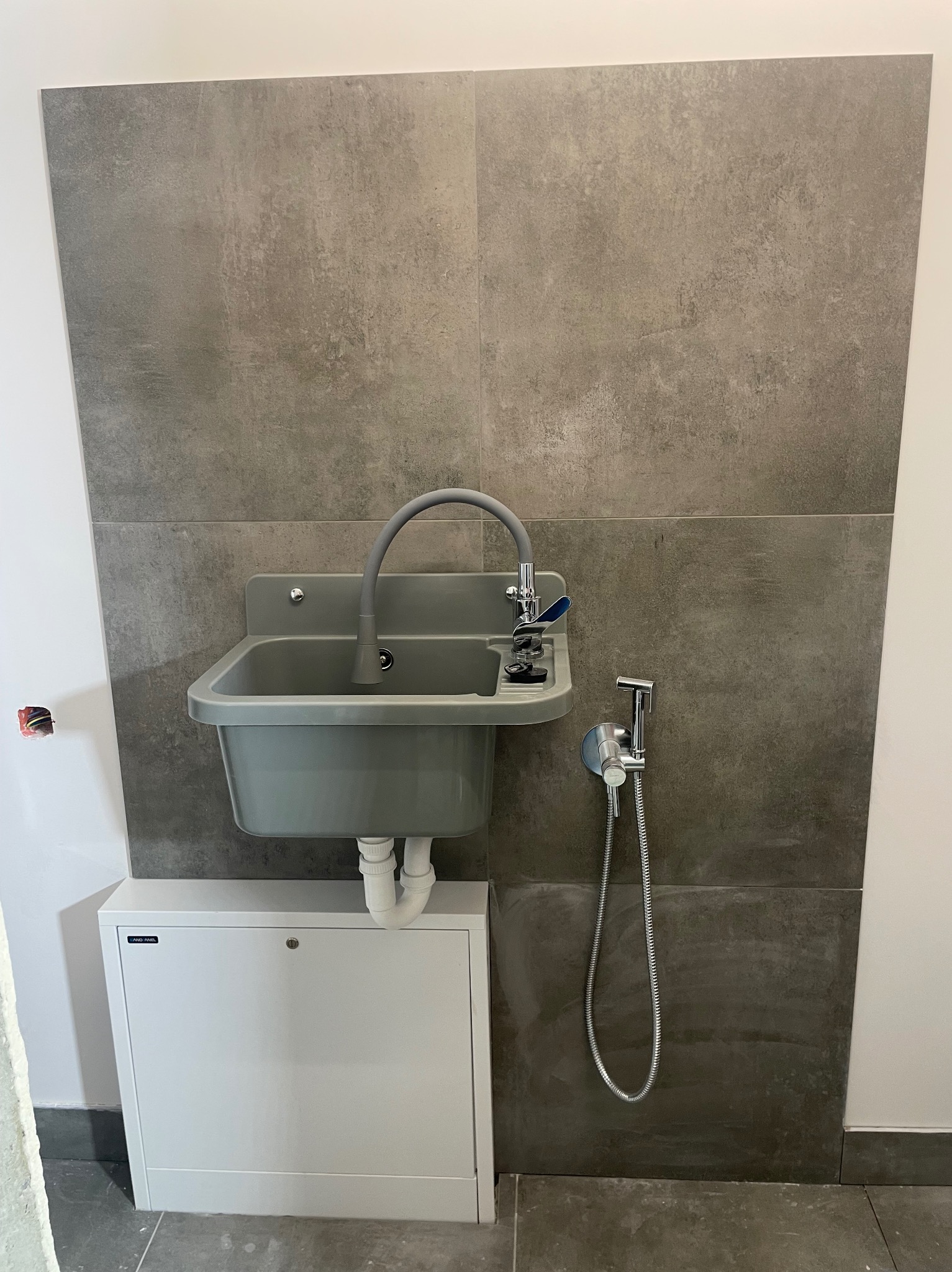 Szara umywalka gospodarcza z baterią i bidetką zamontowana na ścianie wyłożonej szarymi płytkami imitującymi beton, z szafką pod spodem.
