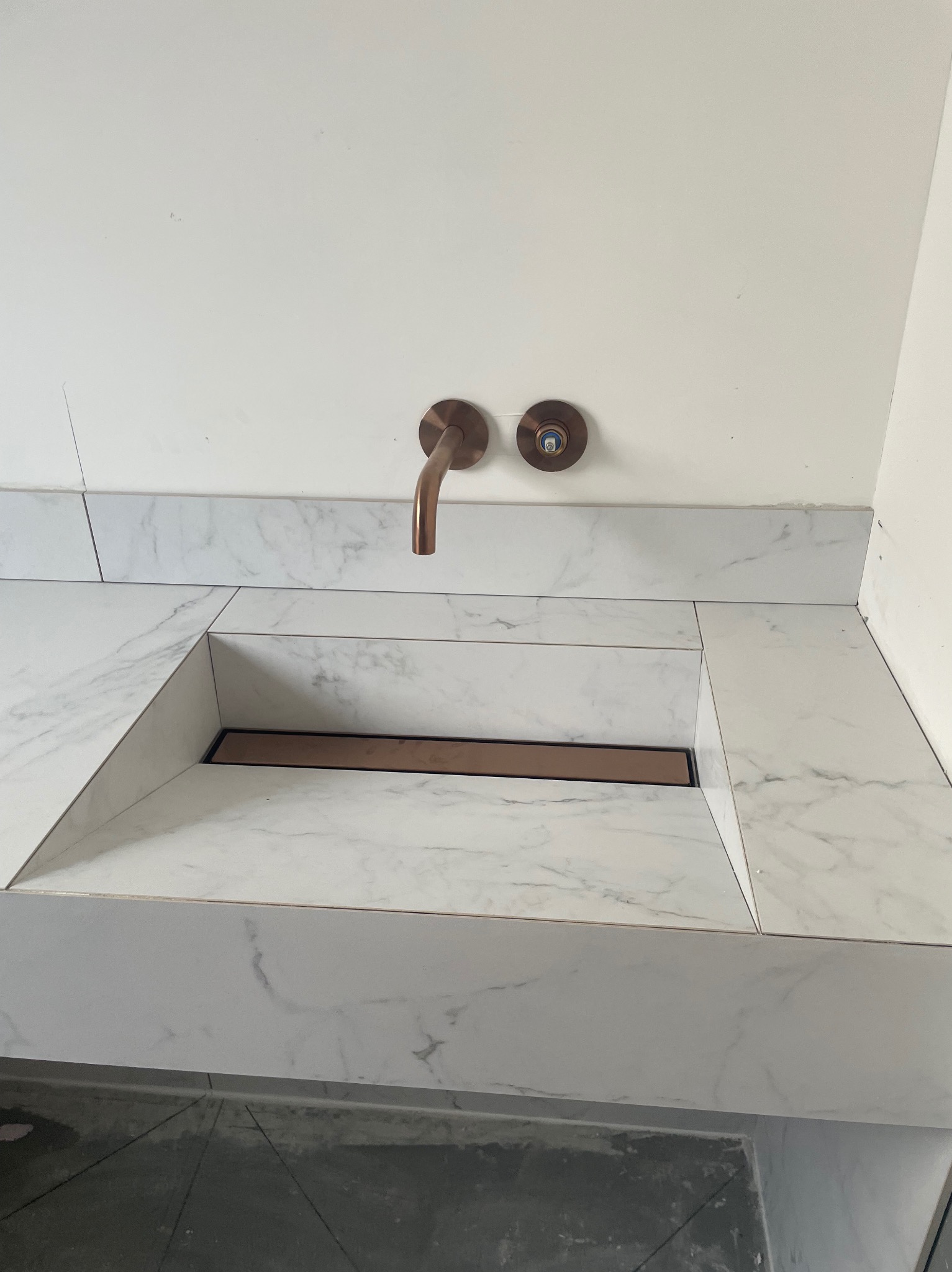 Wpuszczany zlew z odpływem liniowym, obłożony płytkami imitującymi marmur, bateria ścienna w kolorze miedzianym.