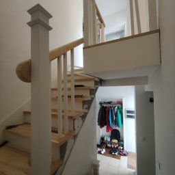JBSM SPÓŁKA Z OGRANICZONĄ ODPOWIEDZIALNOŚCIĄ - Montaż drewnianych schodów z białymi balustradami w jasnym wnętrzu. Widoczne narzędzia i garderoba w tle. Ujęcie z dołu, podkreślające detale wykończenia.