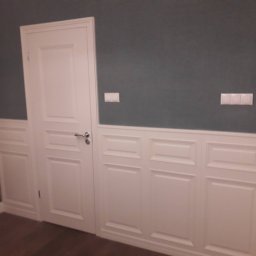 JBSM SPÓŁKA Z OGRANICZONĄ ODPOWIEDZIALNOŚCIĄ - Białe, panelowe boazerie ścienne z drzwiami w stylu angielskim na tle szarej ściany. Klasyczne wykończenie wnętrza z drewnianymi elementami i widocznymi włącznikami światła.