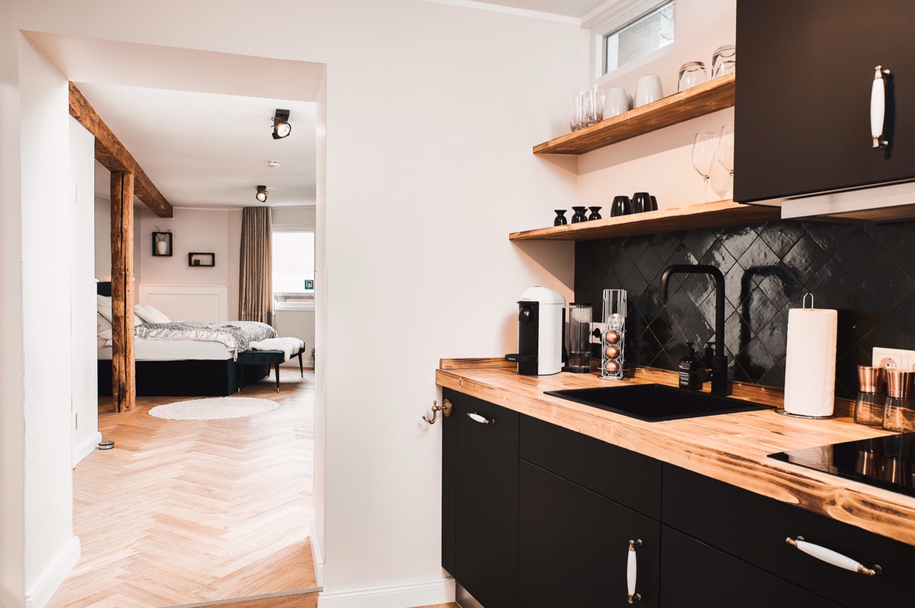 Nowoczesna, czarno-drewniana kuchnia z blatem z litego drewna i czarną armaturą, widok na sypialnię z parkietem w jodełkę i drewnianą belką.