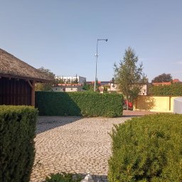 Siwe Ogrody - Widok na posesję z brukowanym podjazdem, równo przyciętym żywopłotem i altaną z trzcinowym dachem w słoneczny dzień.