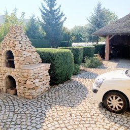 Siwe Ogrody - Ogród z równo przyciętym żywopłotem, kamiennym grillem i brukowanym podjazdem, częściowo zasłoniętym przez biały samochód.