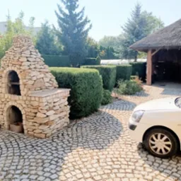 Ogród z równo przyciętym żywopłotem, kamiennym grillem i brukowanym podjazdem, częściowo zasłoniętym przez biały samochód.