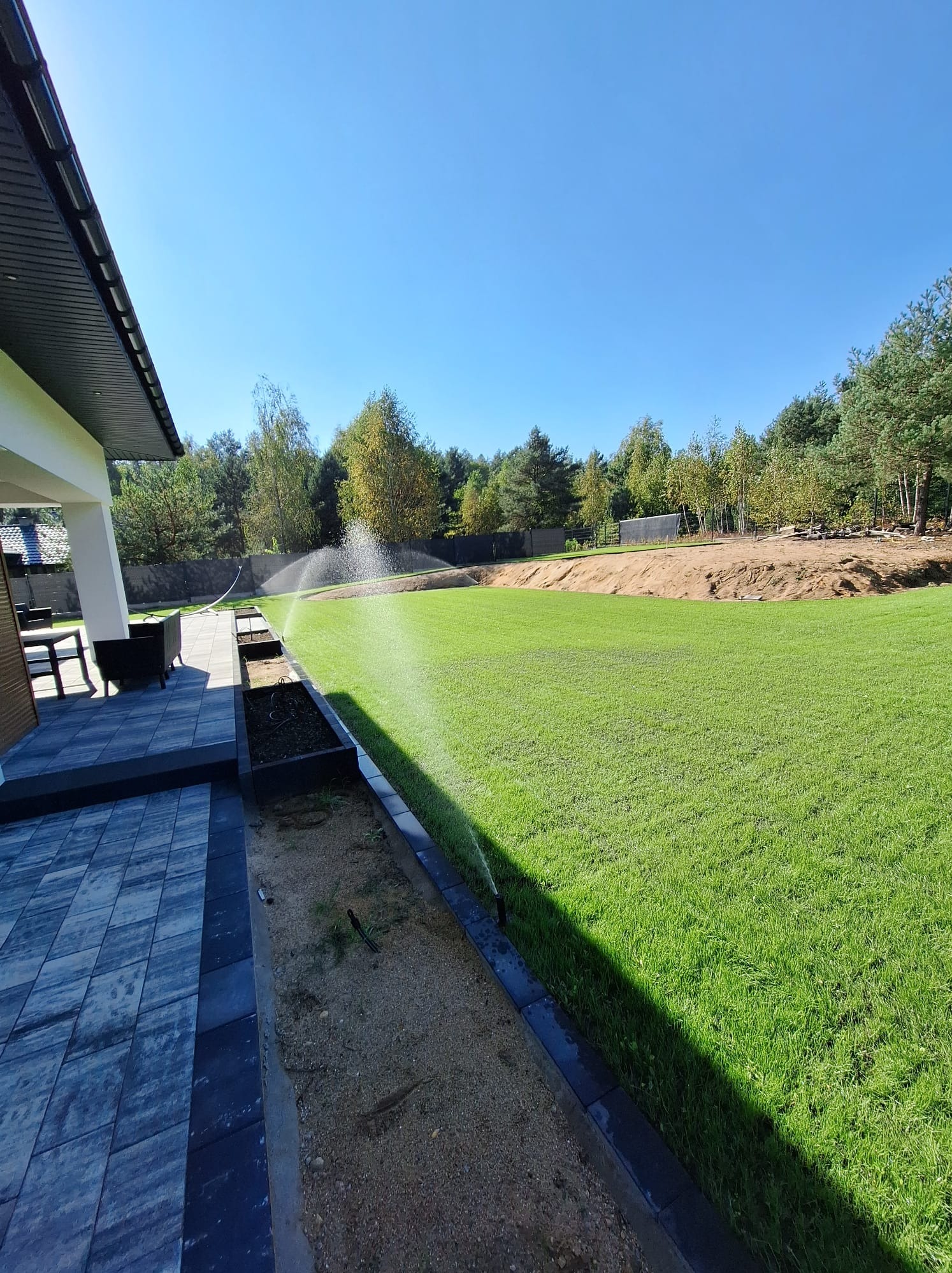 Automatyczne zraszacze nawadniające świeżo założony trawnik z rolki, widok z perspektywy tarasu wyłożonego szarą kostką. W tle widoczny fragment domu z białymi kolumnami i drzewa.
