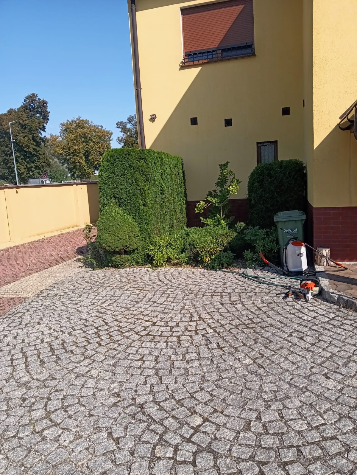 Przycięte, formowane krzewy iglaste przed budynkiem z kostki brukowej, obok stoi opryskiwacz i kosiarka spalinowa.
