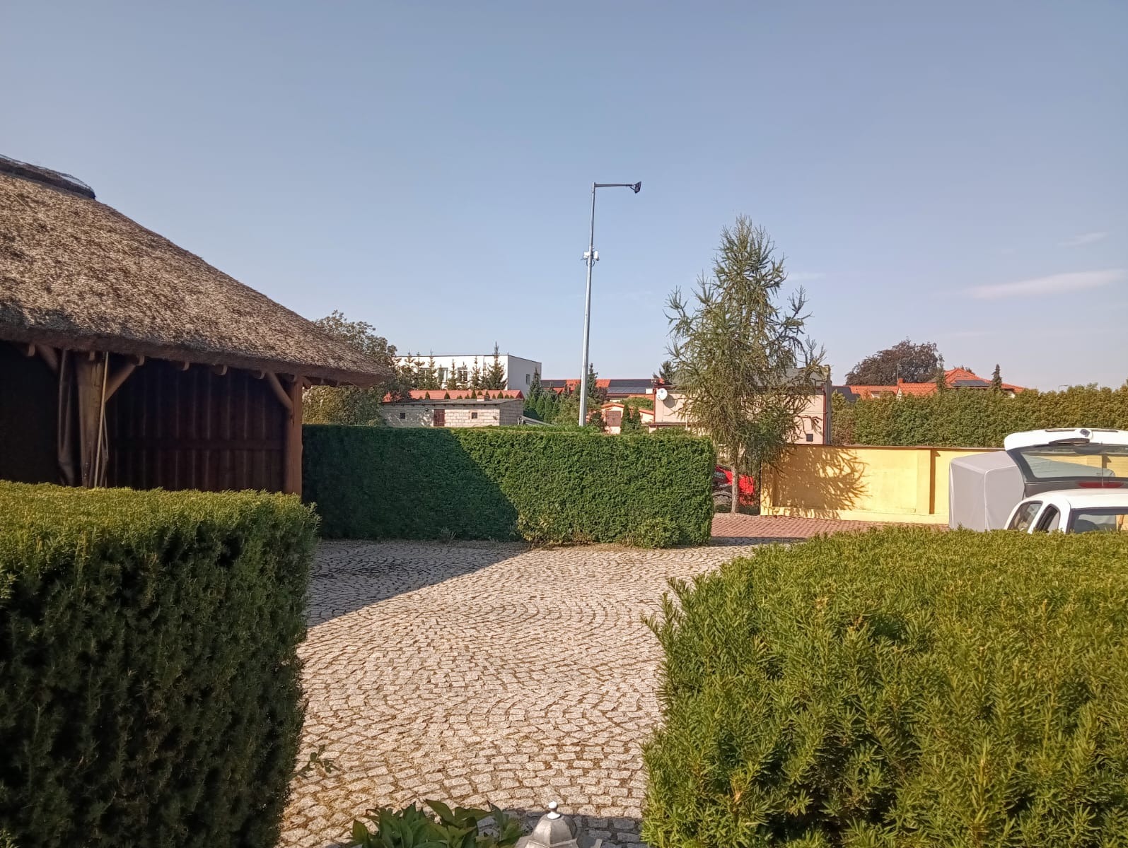 Widok na posesję z brukowanym podjazdem, równo przyciętym żywopłotem i altaną z trzcinowym dachem w słoneczny dzień.