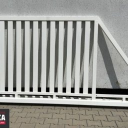 Ogrodzenia.Spawanie - Biały, metalowy fragment bramy z pionowymi szczeblami, oparty o ścianę budynku i kostkę brukową, z widocznym logo producenta konstrukcji stalowych.