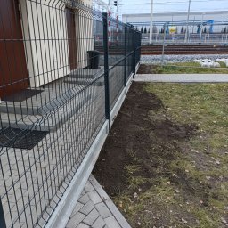 Ogrodzenia.Spawanie - Nowoczesne, szare ogrodzenie panelowe z podmurówką betonową, przylegające do budynku z brązowymi drzwiami, w tle widoczne tory kolejowe i sygnalizacja świetlna.