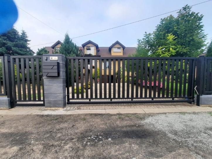 Nowoczesna, przesuwna brama w kolorze antracytu z pionowych profili, zintegrowana ze skrzynką na listy na słupku z szarej kostki, na tle domu z poddaszem.