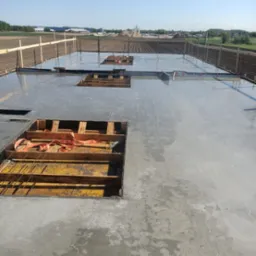 Zalany fundament z widocznym zbrojeniem i drewnianym szalunkiem. W tle pole uprawne i zabudowania.