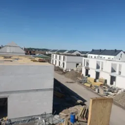 Wysoki kąt na budowę szeregowców z białego bloczka, widoczne ściany i stropy w stanie surowym otwarte, materiały budowlane i droga dojazdowa. Błękitne niebo.