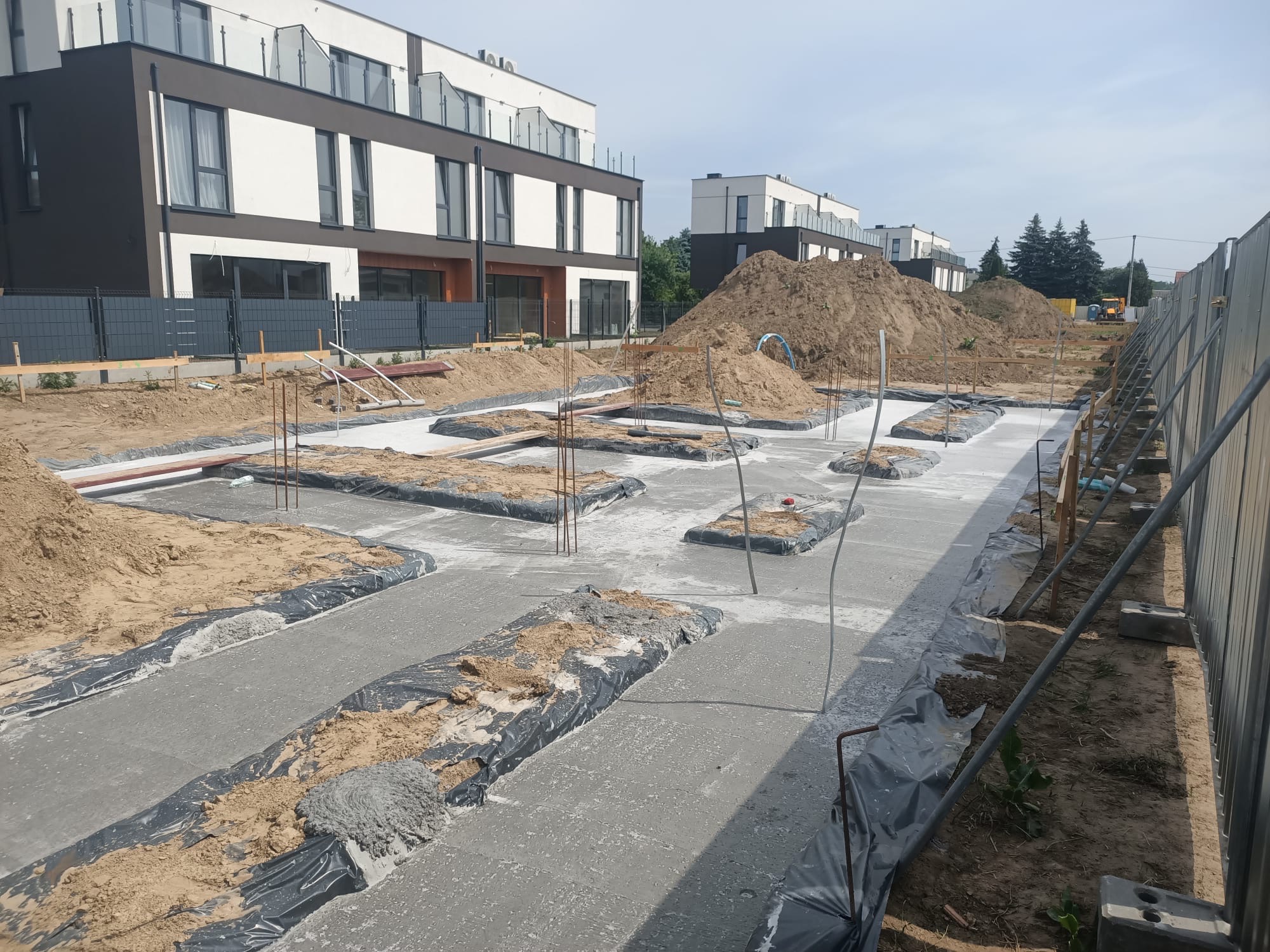 Wylane fundamenty pod budowę domu wielorodzinnego, widoczne zbrojenia i zabezpieczenia folią. W tle budynki w trakcie budowy i hałdy piachu.
