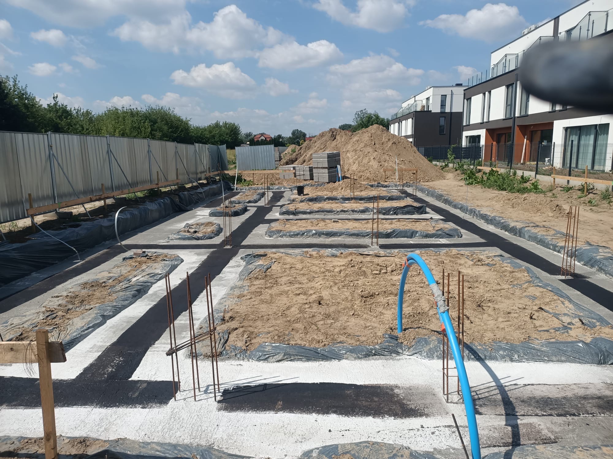 Przygotowany fundament pod budowę domów jednorodzinnych, z widocznym zbrojeniem i izolacją, na tle nowoczesnych budynków i błękitnego nieba.