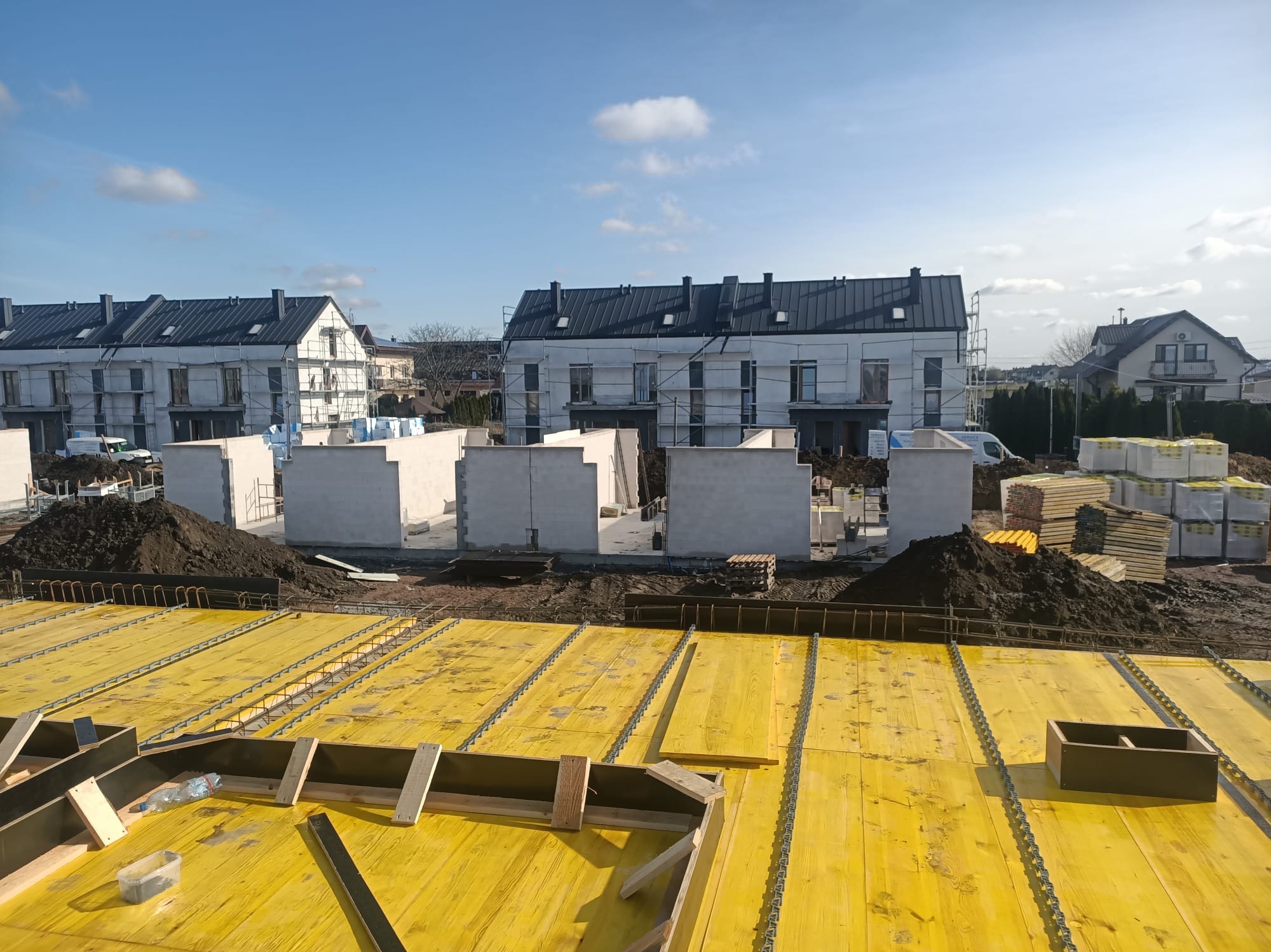 Żółte szalunki stropowe zbrojone stalą na tle wznoszonych ścian domów w zabudowie szeregowej, częściowo w rusztowaniach. Widok z góry, w tle błękitne niebo.